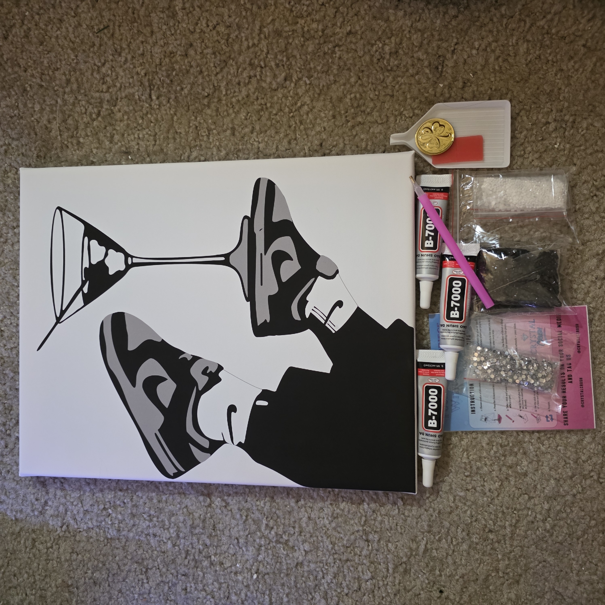 The image shows a diamond painting kit laid out on a carpeted floor. The kit includes a canvas with a black and gray graphic of a person holding a martini glass, two tubes of B-7000 adhesive glue, a pink diamond painting pen, a small plastic tray with a gold shamrock charm and a red adhesive square, and several small plastic bags containing various sizes of rhinestones. The overall scene is presented from a top-down perspective. There is no indication of the specific location within Portland, United States of America.