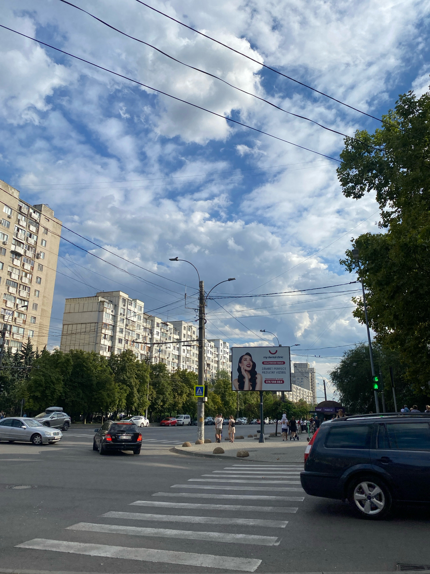 The image captures a street scene in Chișinău, Moldova, likely on a cloudy day. In the foreground, a crosswalk is visible on a paved road. A dark-colored station wagon is parked to the right, partially obscuring the crosswalk. Further back, a black sedan is turning. Several other cars are visible at an intersection, along with pedestrians walking on a sidewalk.
Dominating the background are multi-story apartment buildings, typical of Soviet-era architecture, with numerous windows and balconies. Tall trees line the street, and a billboard for "my dental clinic" with the slogan "HOLLYWOOD SMILE" is displayed prominently. Power lines crisscross the sky, and streetlights and traffic signals are present. The overall impression is a busy urban environment with a mix of residential buildings, traffic, and infrastructure.
