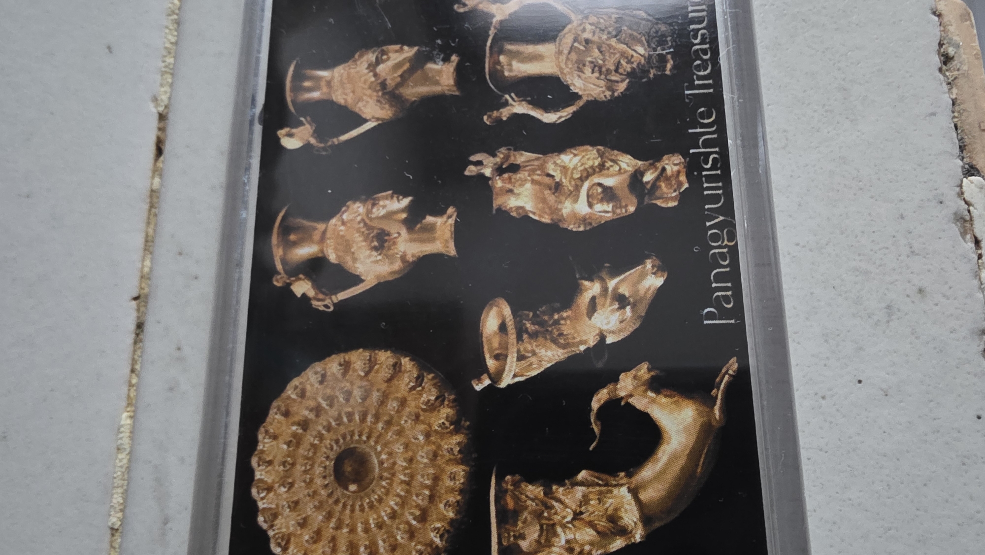 The image is a close-up shot of what appears to be a postcard or photograph encased in a clear plastic sleeve. The postcard features a collection of golden artifacts displayed against a black background. The objects include several rhytons (drinking horns) shaped like animal heads (rams, horses, and others) and a shallow dish with intricate concentric patterns. The text "Panagyurishte Treasure" is visible in bold, upright letters on the right side of the image. The casing shows signs of wear and is resting on a white or light-colored surface. There is no indication of location in the city of Blagoevgrad, Bulgaria from this image.
