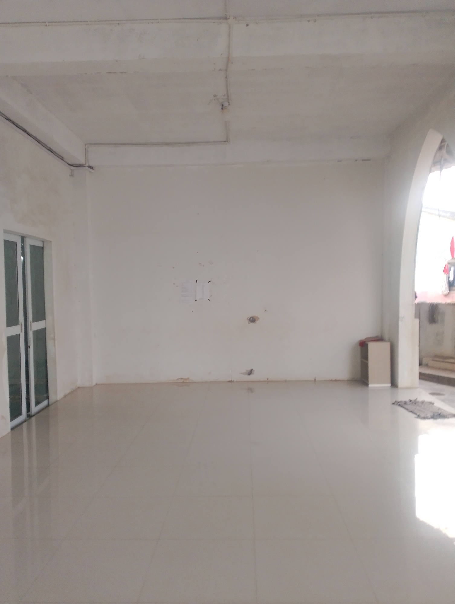 The image depicts an interior view of a spacious, unfinished room, likely a commercial or residential space, located in Batam, Indonesia. The room features white painted walls and a light-colored tiled floor, which reflects the ambient light. The ceiling is also white, with visible electrical conduits and some staining.
On the left side of the room, there is a set of double glass doors with white frames, leading outside. The doors are closed.
The walls are largely bare, with some faint marks and a few pieces of paper attached to the wall in the middle-left section. A small, light-colored cabinet or shelf unit is visible against the wall on the right side, near an arched doorway. Through the archway, a glimpse of an outdoor area can be seen, with what appears to be clothing or laundry hanging and stairs leading upwards. A small rug is placed on the floor in front of the arched opening.
The lighting in the room is bright, with a strong reflection coming from the floor on the right side, suggesting a light source from outside. The overall impression is one of a newly constructed or renovated space that is yet to be furnished or fully completed. The location in Batam, Indonesia, suggests a tropical environment, which might influence the materials and design of the building.