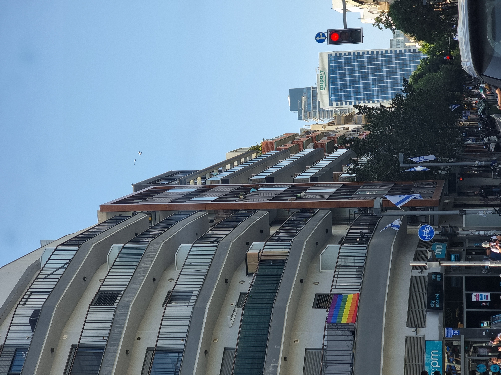 The image depicts a street scene in Tel Aviv, Israel, featuring a modern, multi-story building with curved balconies. A rainbow flag hangs from one of the balconies. Several Israeli flags are visible along the street. Street signs and a bicycle lane sign are also present. A traffic light is visible. In the background, a taller building is identified with the Hebrew word "דיסקונט" (Discount). People are seen on the sidewalk and in the street. The sky is clear and blue.