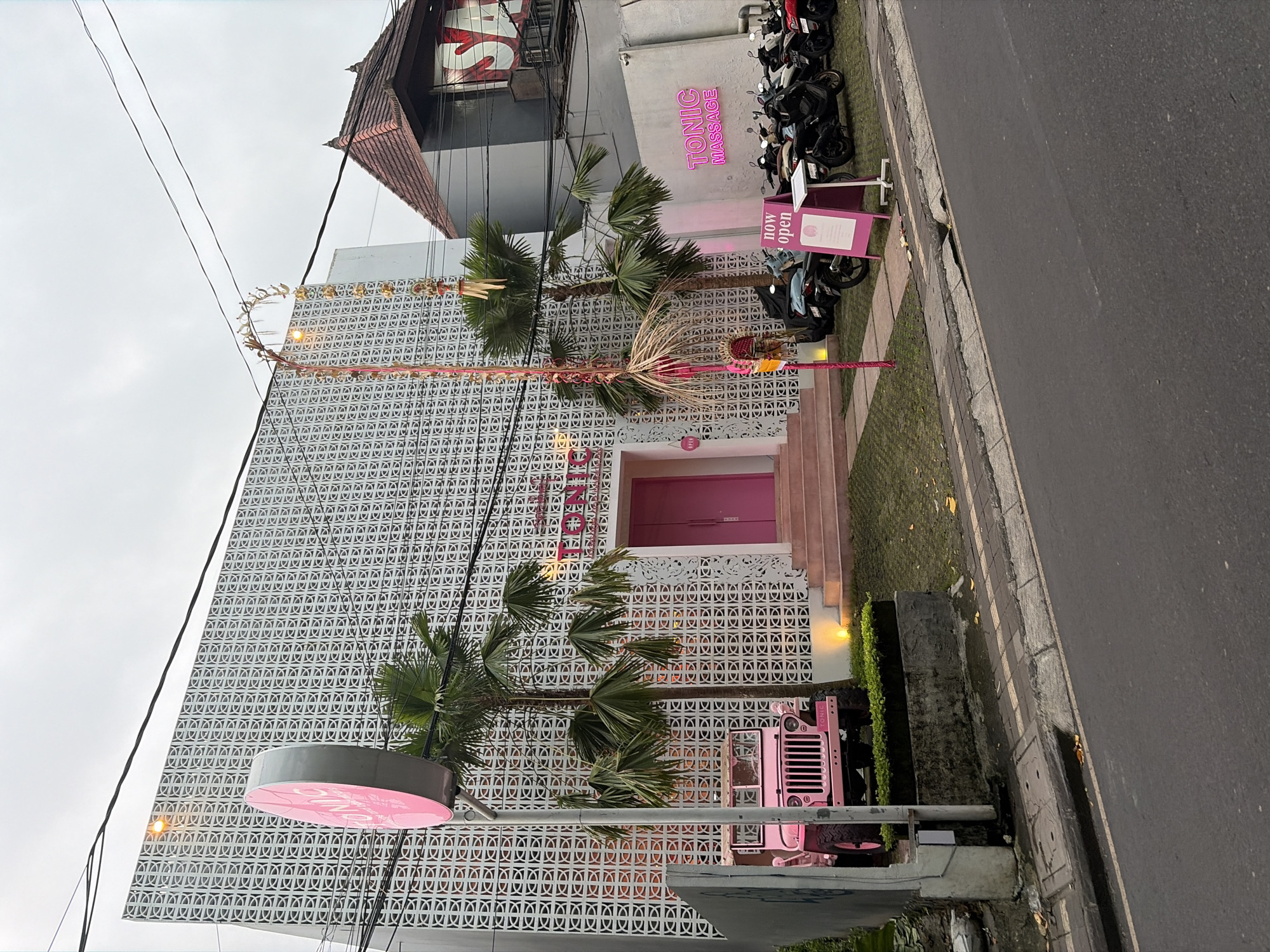 The image depicts the exterior of a business establishment in Ubud District, Indonesia, likely a massage parlor, given the signage. The building features a distinctive white patterned facade with geometric cutouts. In front of the building, a bright pink jeep is parked, adding a vibrant splash of color to the scene. Several palm trees are strategically placed around the entrance, contributing to a tropical ambiance. A pink circular sign with the business's name is visible to the left, while another neon pink sign on the building reads "TONIC MASSAGE." A smaller pink sign on the right reads "now open." A row of motorcycles is parked to the right of the entrance, suggesting a local business catering to customers with transportation. The overall environment appears to be a street scene, with a road and sidewalk in the foreground, and power lines overhead. The sky is overcast, indicating a potentially cloudy or rainy day.