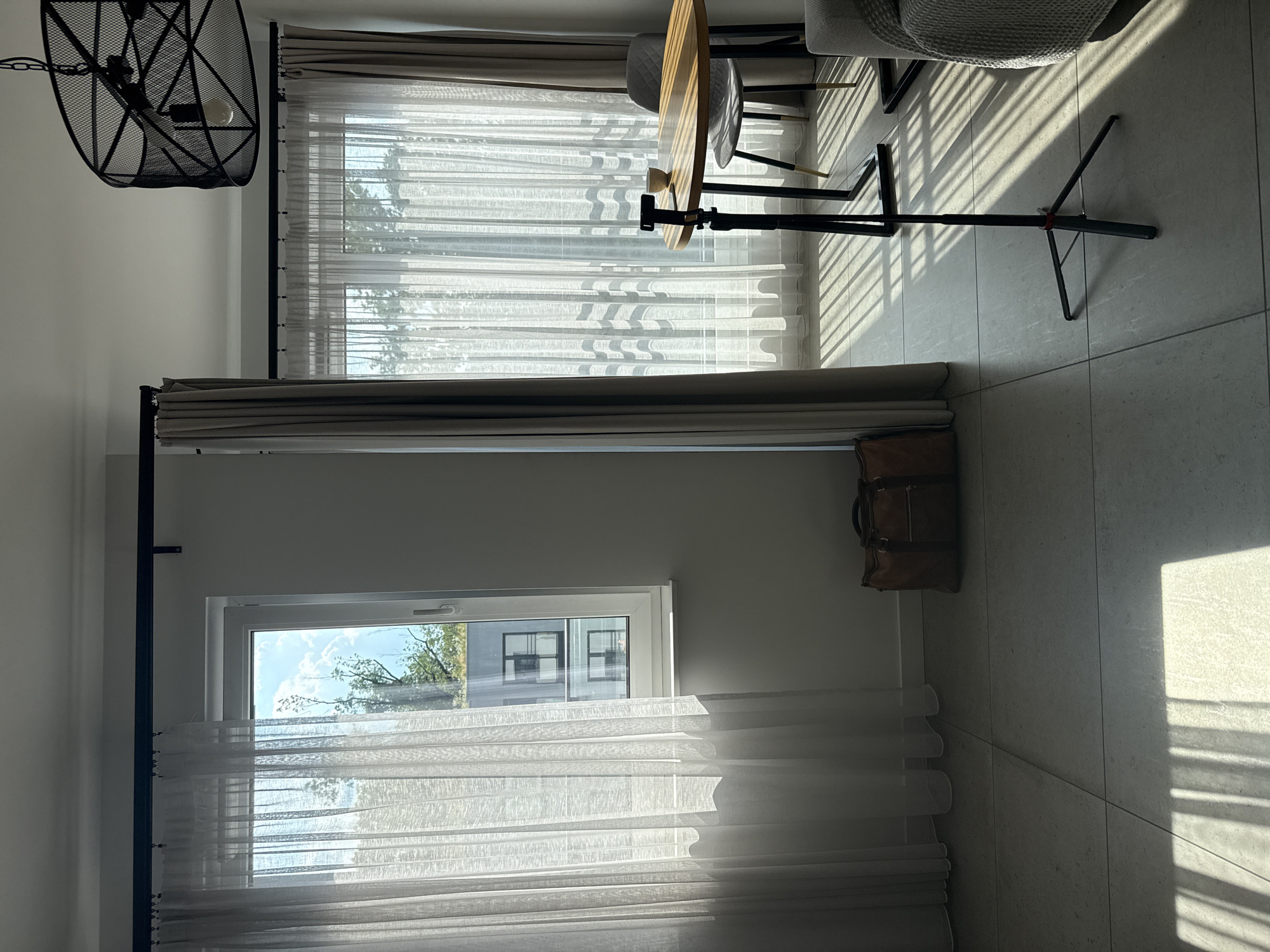 The image depicts an interior scene bathed in sunlight streaming through sheer curtains. On the left, a window overlooks an urban environment with modern buildings. A brown leather duffel bag rests on the tiled floor near the window. In the center right, a round wooden table is set with a small bowl and a smartphone on a tripod. A gray upholstered chair is pulled up to the table. Further to the right, a portion of a sofa with a textured gray throw is visible. A modern black pendant light fixture hangs from the ceiling. The scene suggests a living area or a study, with an emphasis on natural light and minimalist decor. The location is identified as Unknown, Unknown.