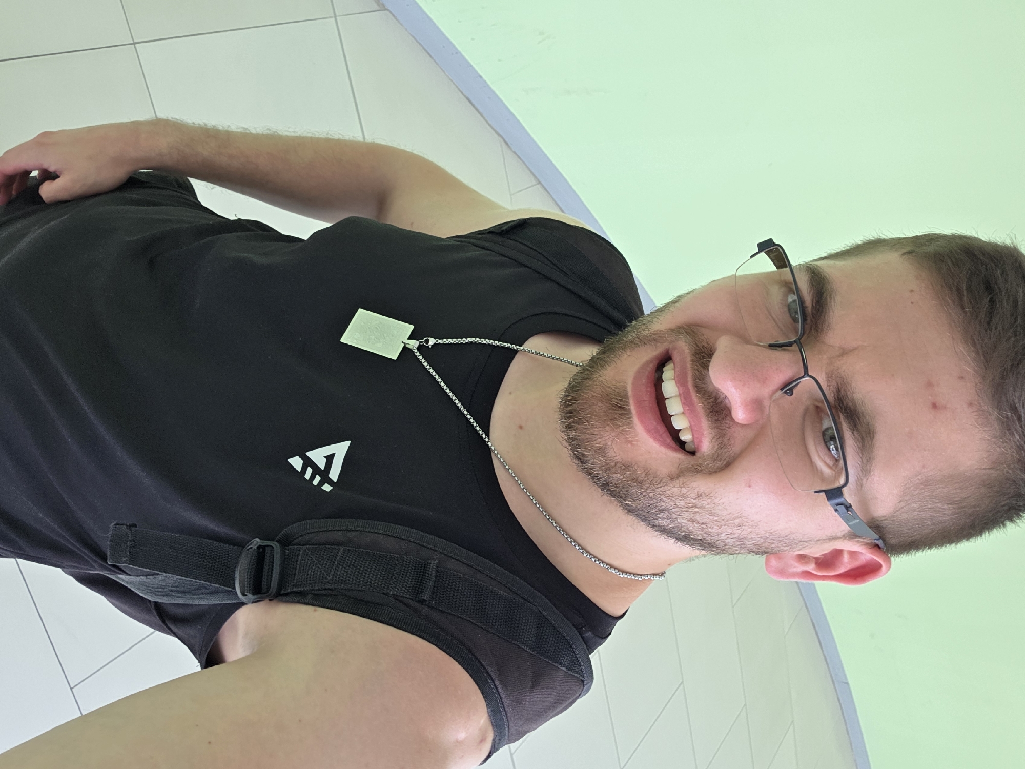 A medium shot depicts a man with brown hair and a beard, wearing glasses and a black shirt. He is wearing a silver necklace with a tag. His facial expression appears to be a slight smile. He is indoors, standing on a white tiled floor with a pale green wall in the background. The location is unknown but the man is in Plovdiv, Bulgaria.
