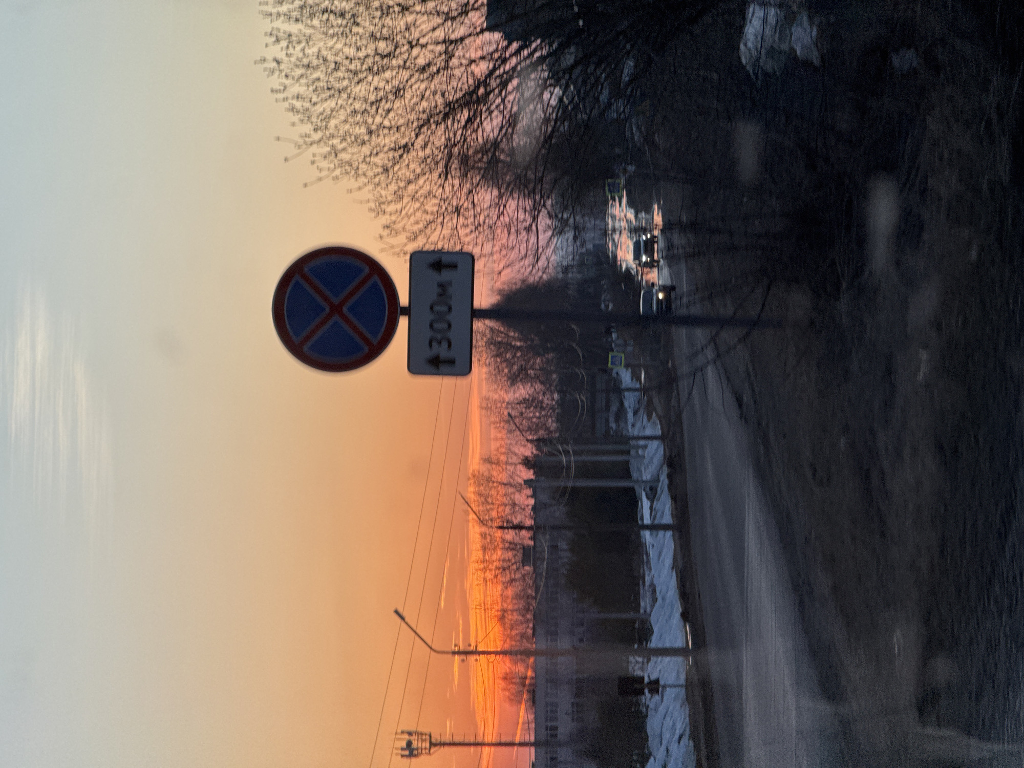 The image depicts a street scene in Прикамский, Russia, at sunset. A road runs through the frame from the foreground to the distance. On the left side of the road are snow banks, and on the right is a curb. The sky is a gradient of orange and pink, indicating a setting sun. In the foreground, there is a road sign with a blue circle and a red "X" indicating no parking, next to a sign that says 300M with an arrow pointing up. In the distance, there are vehicles traveling on the road. Bare trees are visible in the middle of the image, and street lights are scattered along the road. Buildings are visible in the distance under the sunset.