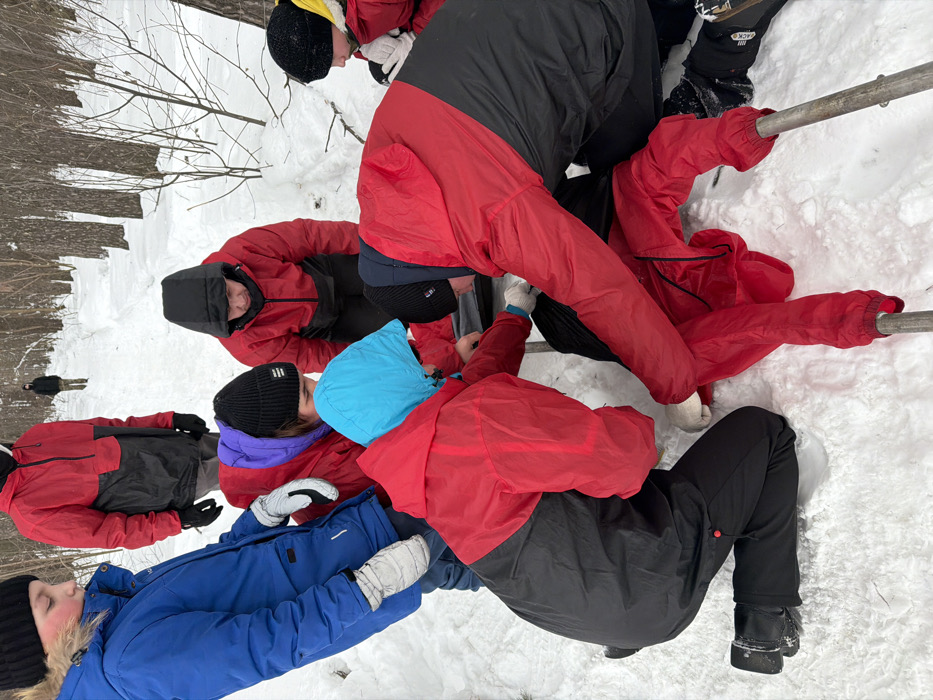 Several individuals in snow gear huddle together outdoors on a snowy surface near a wooded area. They wear jackets in various colors, including red, black, blue, and purple, and hats. Some are interacting with a metal railing. The composition is from a high-angle perspective, capturing the group's close proximity and activity in the snowy environment. Two figures stand distantly in the background.