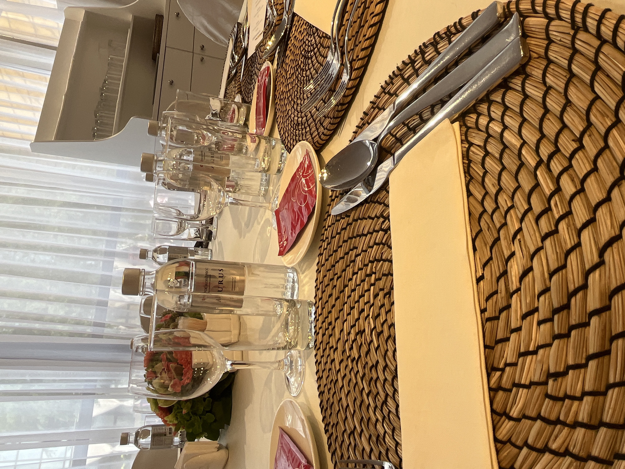 This image captures a finely set dining table indoors, likely prepared for a formal event or meal in Shushary, Russia. The scene is illuminated by natural daylight filtering through a large window draped with sheer white curtains, visible in the background on the left.
The table is covered with a pristine white tablecloth, upon which multiple place settings are arranged. Each setting features a distinctive round placemat crafted from woven natural fibers, topped with a small beige plate. A folded red napkin, possibly with a decorative pattern, rests on each plate. Stainless steel silverware, including forks, knives, and spoons, is neatly positioned beside the plates.
Numerous clear glassware items are present, including elegant wine glasses and smaller water glasses. Several small, clear bottles of water are also lined up on the table, bearing visible text that reads "AURUS" and "РОССИЙСКОЕ ПРЕСС" (Russian Press). Some of the glasses contain small, vibrant floral arrangements with red flowers and green foliage, adding a decorative touch.
In the upper left background, a white cabinet or shelving unit with drawers and small compartments is visible, appearing to store more glassware or dining essentials. No people are present in the image, suggesting the table is either awaiting guests or has just been cleared. The overall setting conveys an atmosphere of prepared elegance and order, indicative of a special occasion taking place during the daytime.
