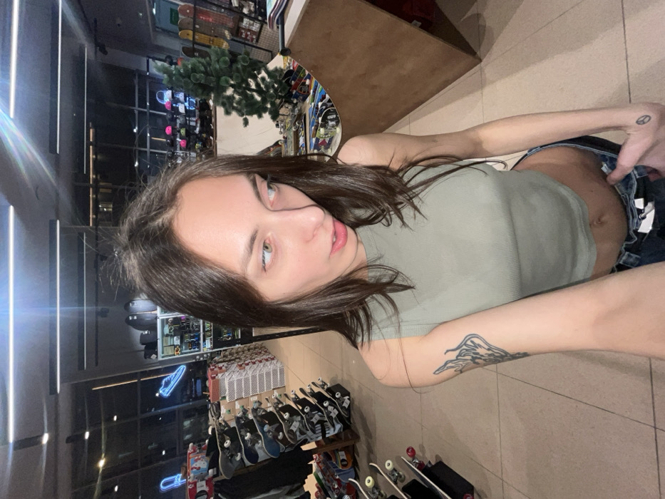 A selfie-style image displays a young individual with long, dark brown hair, light-colored eyes, and fair skin. The subject is wearing a light olive-green ribbed crop top, exposing the midriff, and dark blue jeans. A black tattoo depicting a flame-like motif is visible on the upper left arm, and a small black peace symbol tattoo is present on the back of the left hand. The individual is looking towards the viewer's left, with the mouth slightly open.
The scene is an indoor retail environment identified as a skate shop in Krasnoyarsk, Russia. The flooring is composed of light-colored tiles, and the ceiling features bright, linear LED strip lighting. To the left of the subject, multiple skateboards are mounted vertically on display racks. A small, artificial green Christmas tree stands in the mid-ground to the right. A wooden mini ramp or quarter pipe is visible on the far right. Additional merchandise, including helmets and bags, is arranged on shelves in the background. A blue neon sign in the shape of a sneaker is visible on the left wall. Large windows in the background show a dark exterior, indicating an evening or nighttime setting. No other individuals are present in the frame.