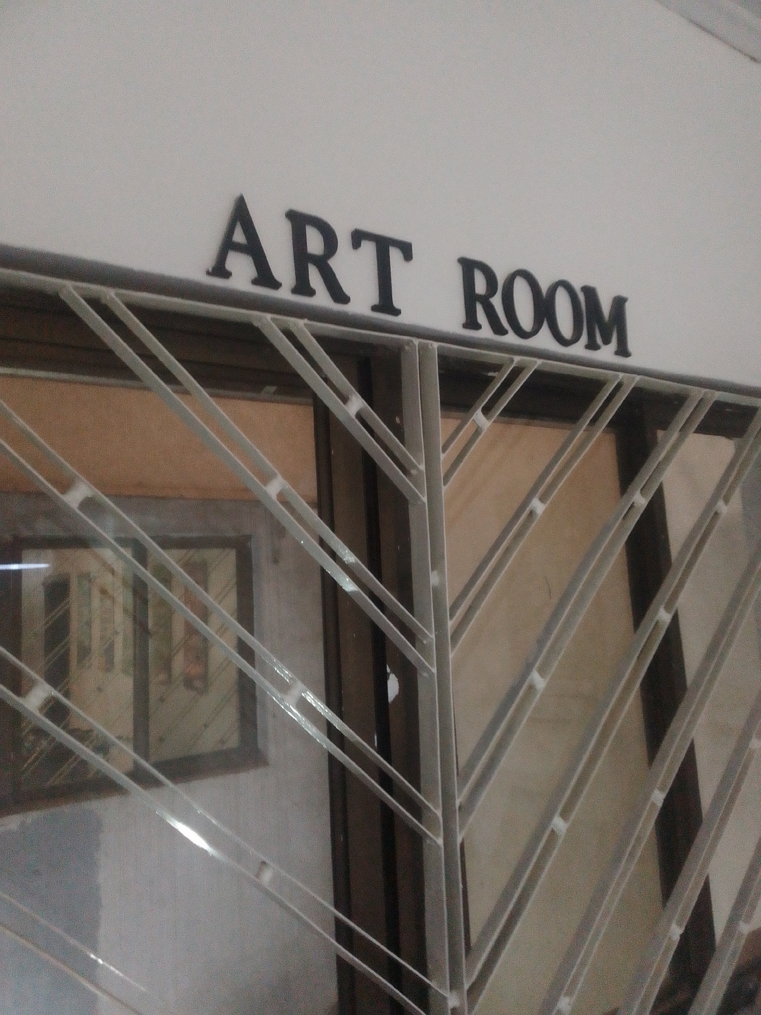A low-angle, eye-level photograph captures the entrance to an "ART ROOM" in Abuja, Nigeria. The words "ART ROOM" are displayed in prominent black, raised capital letters on a stark white wall above a recessed doorway. A light grey or white metal security grille, featuring a diagonal, V-shaped repeating pattern, covers the glass door or window directly below the signage. This grille is secured into dark brown door or window frames. Through the glass and the grille, a light brown or beige interior wall is visible. A framed rectangular object, likely artwork, hangs on this interior wall and is partially discernible behind the grille. Reflections of an interior light source or exterior light are visible on the glass and the metal grille bars. No individuals are present in the scene.