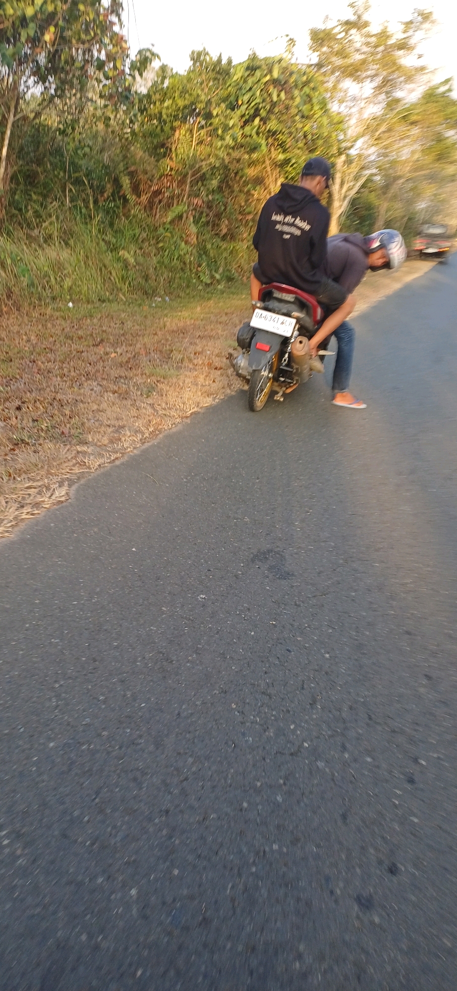 Two individuals are on a red motorcycle on a paved road in Kersik Putih, Indonesia. The person in the back is wearing a black hoodie and a dark baseball cap. The person in the front is wearing a dark hooded shirt and a helmet. The motorcycle has a license plate that reads "DA-6341-LEH". The road is lined with dry grass and trees. The scene appears to be outdoors during daylight.