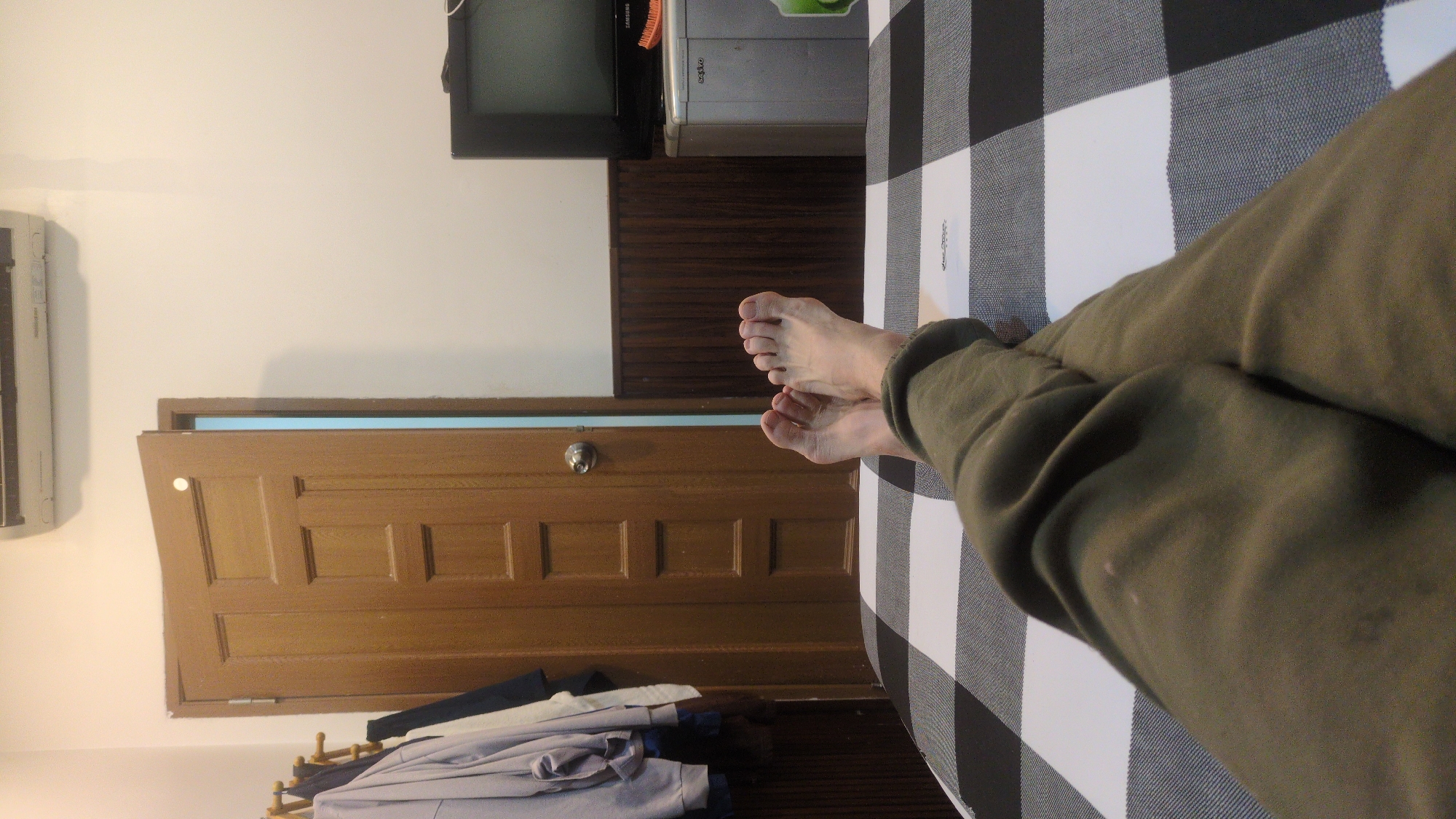 A photograph taken indoors in Phan Rang – Tháp Chàm, Vietnam depicts a pair of bare feet resting on a black and white checkered surface, possibly a bed or blanket. The legs are clad in olive-colored trousers. In the background, a wooden door is slightly ajar, revealing a glimpse of blue light. Adjacent to the door is an air conditioning unit mounted on the wall. Above the door, a retro television set sits atop a metal cabinet. A pile of clothing is visible near the base of the door. The wall behind the television and cabinet is painted white, with a section of wooden paneling below.