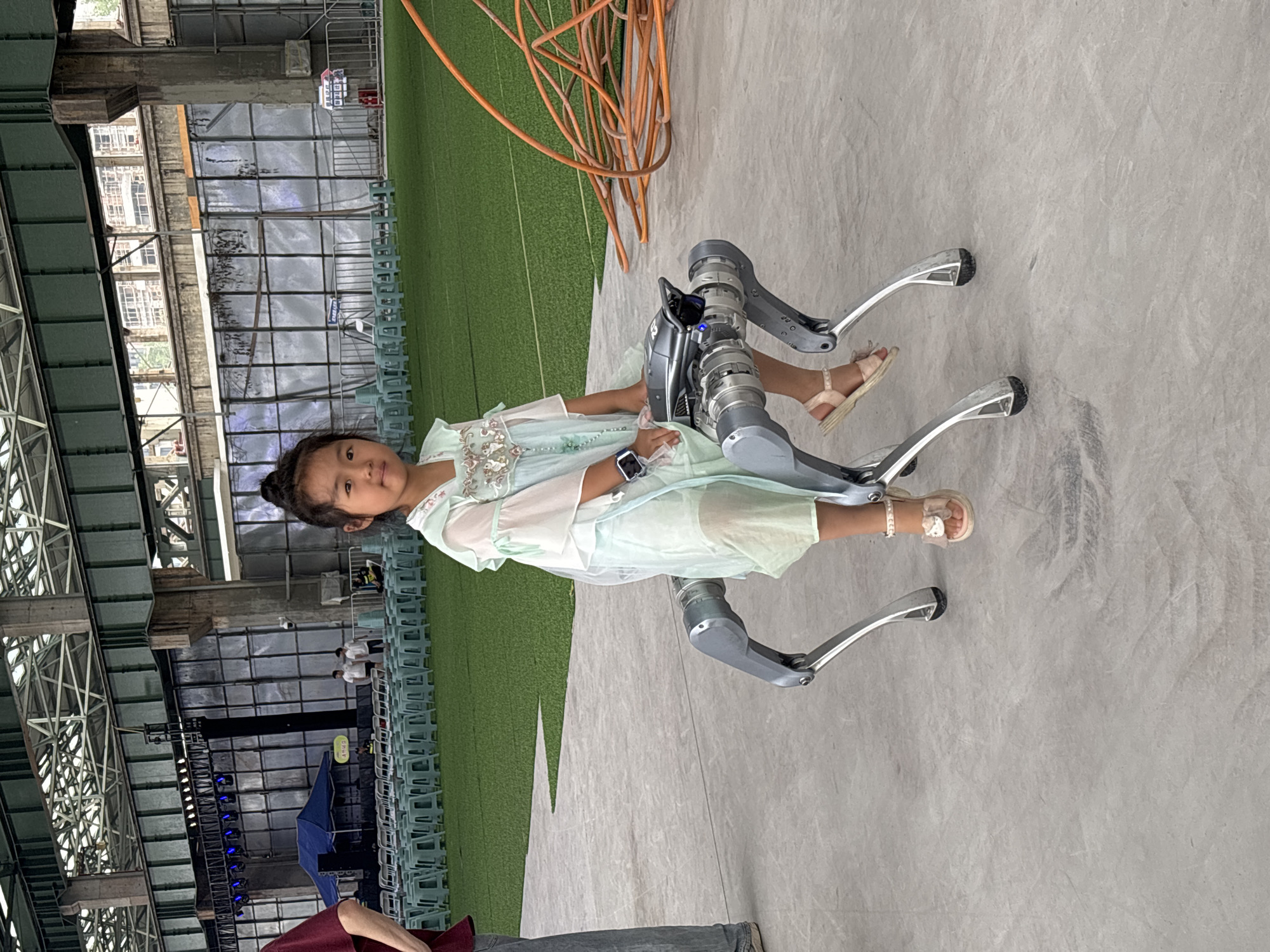 In the city of Dadukou, China, an East Asian girl with dark hair pulled into a bun sits on a robotic quadruped. She is wearing a pale blue dress with white floral embroidery on the bodice and sheer ruffled sleeves, and her feet are adorned with pink sandals featuring bow accents and pearls. The robotic dog, silver and gray, has four articulated legs and a sleek, metallic body with a dark top and a blue light illuminated on its head. The girl is looking towards the camera, with a slight smile.
The scene appears to be an indoor event space, possibly a convention center or exhibition hall. The floor is a smooth, light gray concrete. In the background, rows of light blue plastic chairs are neatly arranged on a green turf surface, suggesting a waiting area or seating for an audience. Behind the chairs, a large stage with a black backdrop and a blue tent are visible, along with a light rig and speakers, indicating a performance or presentation area. Large concrete pillars and a metallic ceiling structure are prominent features of the architecture. To the right of the robotic dog, a large coil of orange cabling is present on the concrete floor. The overall impression is of a modern, open space set up for an event.