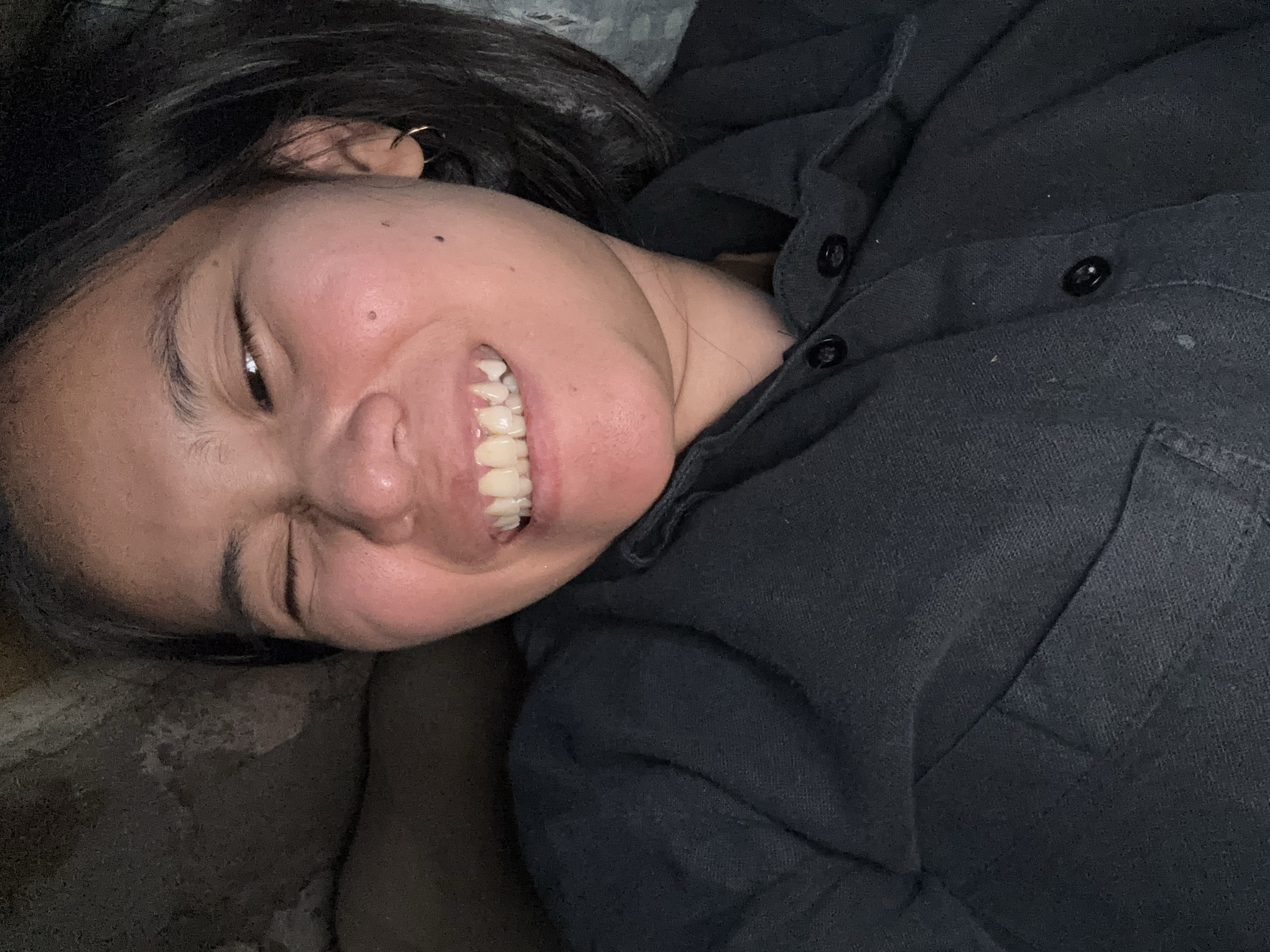 The image is a close-up, low-angle, selfie-style photograph of a young woman. She has dark hair, dark eyes, and her mouth is open in a wide smile, revealing her teeth. One eye is partially closed. She is wearing a dark collared shirt with buttons. The background is dark and appears to be a textured surface, possibly a bed or sofa, with a hint of a patterned fabric on the right side of the frame. The lighting is dim, casting shadows and highlighting her facial features. The image was taken in East Jakarta, Indonesia.