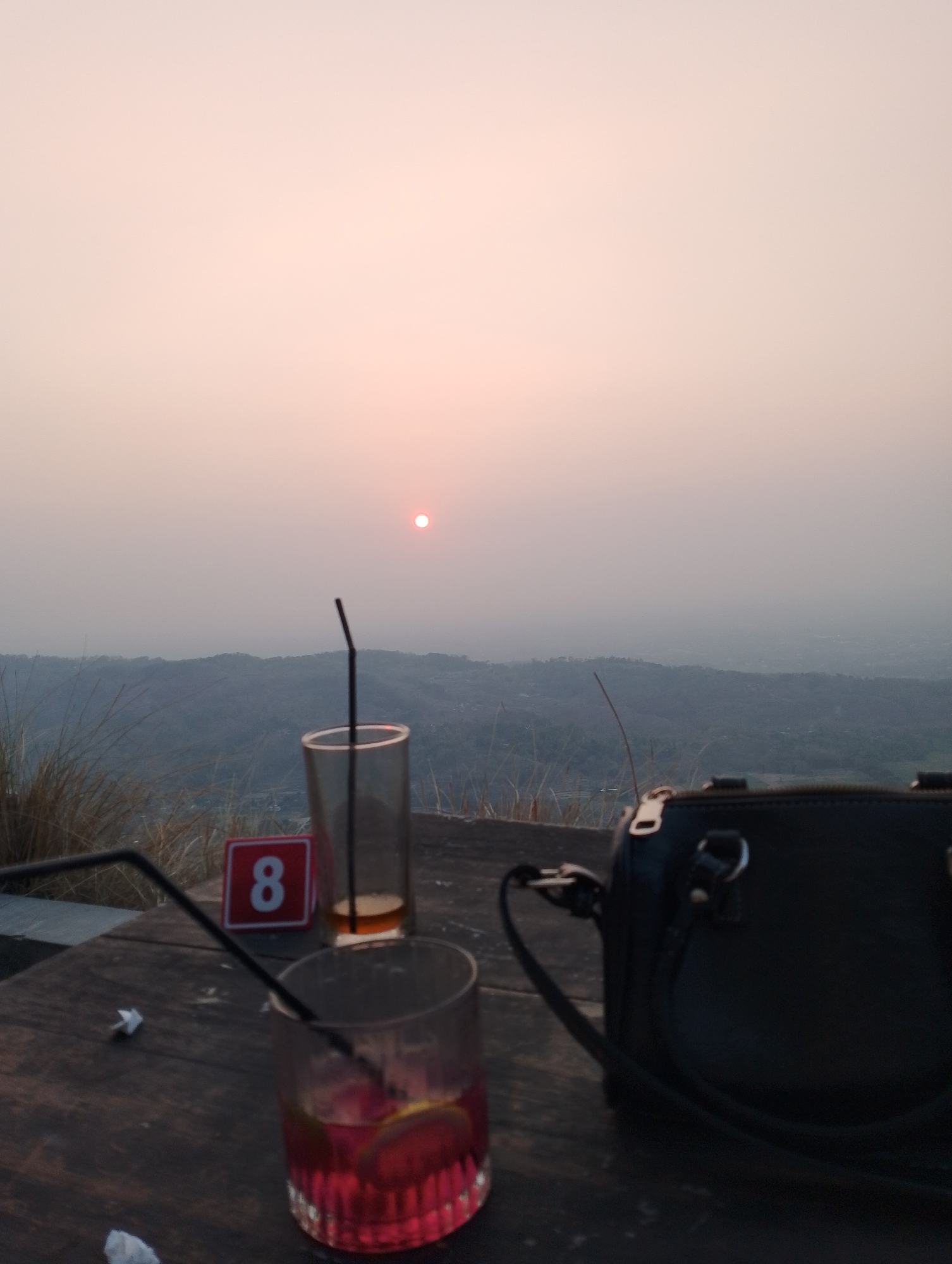 The image depicts a sunset scene from a high vantage point overlooking a hazy landscape, likely Srimulyo, Indonesia, given the context. In the foreground, a wooden table holds two drinks. The drink on the left is a red beverage in a glass with ice and lemon slices, adorned with a black straw. To its right, a partially consumed amber-colored drink sits in a taller glass, also with a black straw. Behind these drinks, a small red sign with a white number "8" is visible. A black handbag with gold hardware rests on the table to the right of the frame. The background showcases rolling hills shrouded in mist and a soft, pale sky where a small, circular sun is visible. Tall, dry grass is present on the left side of the image, adding a natural element to the setting. The overall atmosphere suggests a serene, outdoor dining or relaxation spot.