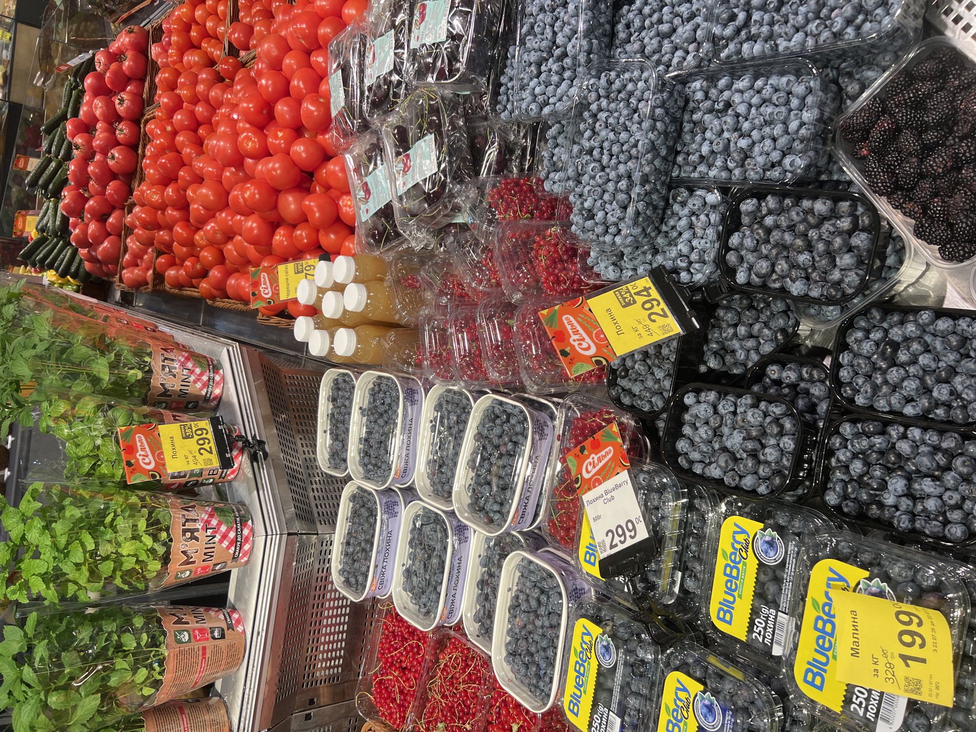 The image shows a produce aisle in a grocery store, likely in Odesa, Ukraine. The display features a variety of fresh fruits and vegetables. In the foreground, there are several containers of blueberries and raspberries, with price tags indicating their cost and weight. To the left, there are potted mint plants for sale. In the middle ground, a large selection of tomatoes is visible, as well as some cucumbers in the background. There are also plastic bottles of juice or other beverages on display. The overall impression is one of a well-stocked and vibrant market.