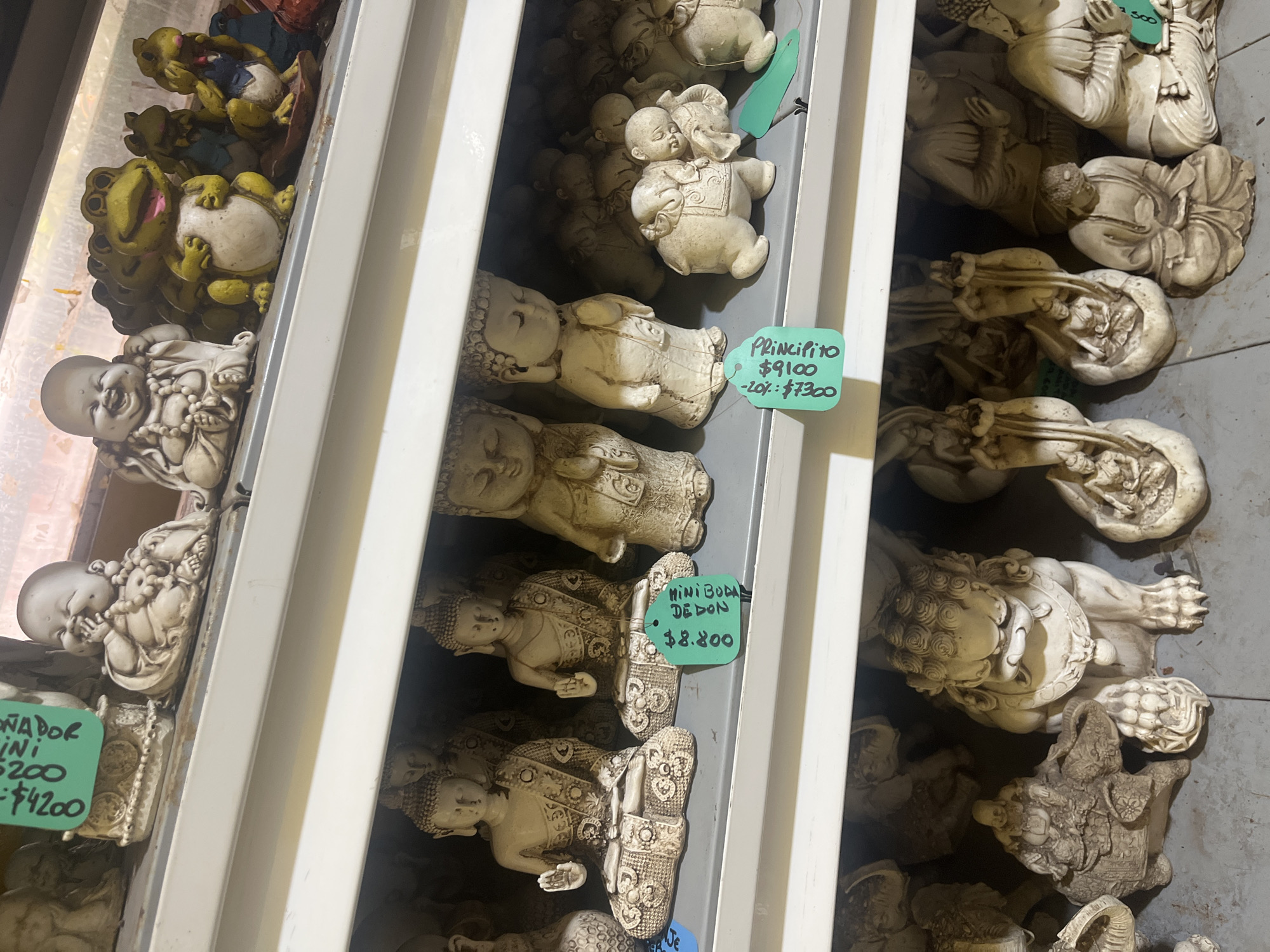 The image shows a collection of small figurines displayed on shelves. The figurines appear to be made of a light-colored material, possibly plaster or resin. They depict various subjects, including Buddhas, smiling children, frogs, and lions. Each figurine is labeled with a price tag. One figurine is labelled "PRINCIPITO" and is priced $9100, with a 20% discount of $7300. Another is labelled "MINI BODA DE DON" and priced at $8800. One label has the text "ONADOR INI" and "200" and "$4200". The image was taken in Buenos Aires, Argentina.