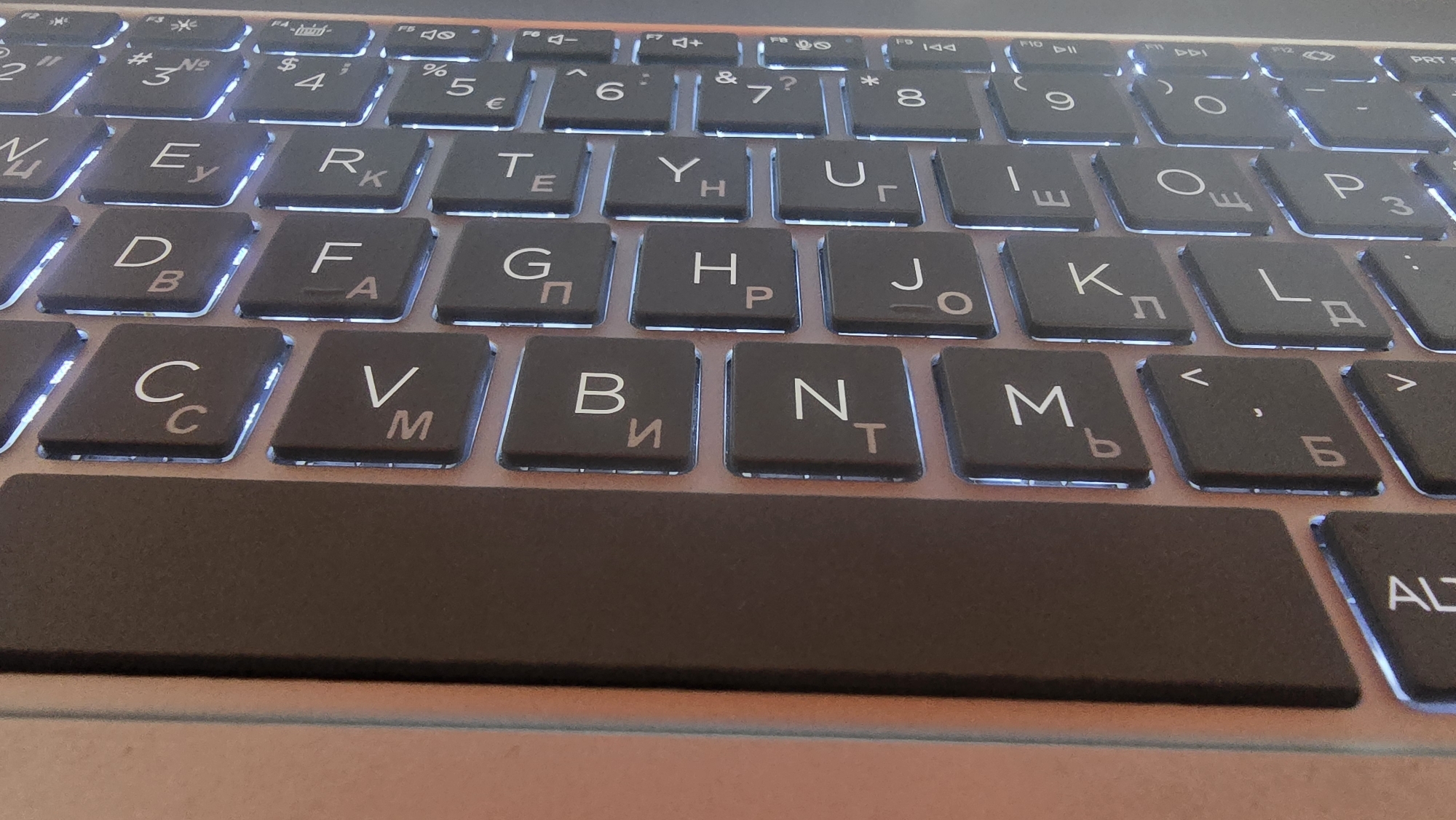 The image shows a close-up view of a backlit keyboard. The keys are dark with illuminated letters and symbols. Several keys are visible, including ones with Cyrillic letters such as 'Е', 'Р', 'Т', 'У', 'Ц', 'О', 'П', 'Л', 'Д', 'К', 'М', 'Н', 'В', 'Б', 'С', 'F', 'G', 'H', 'J', 'D', 'C', and 'V'. There are also number keys and punctuation symbols visible. The keyboard is on a metallic surface, possibly a laptop. The lighting suggests it is either night or a dimly lit room. There is no discernible city, location, or any contextual information that would place it in Ghafurov, Tajikistan.