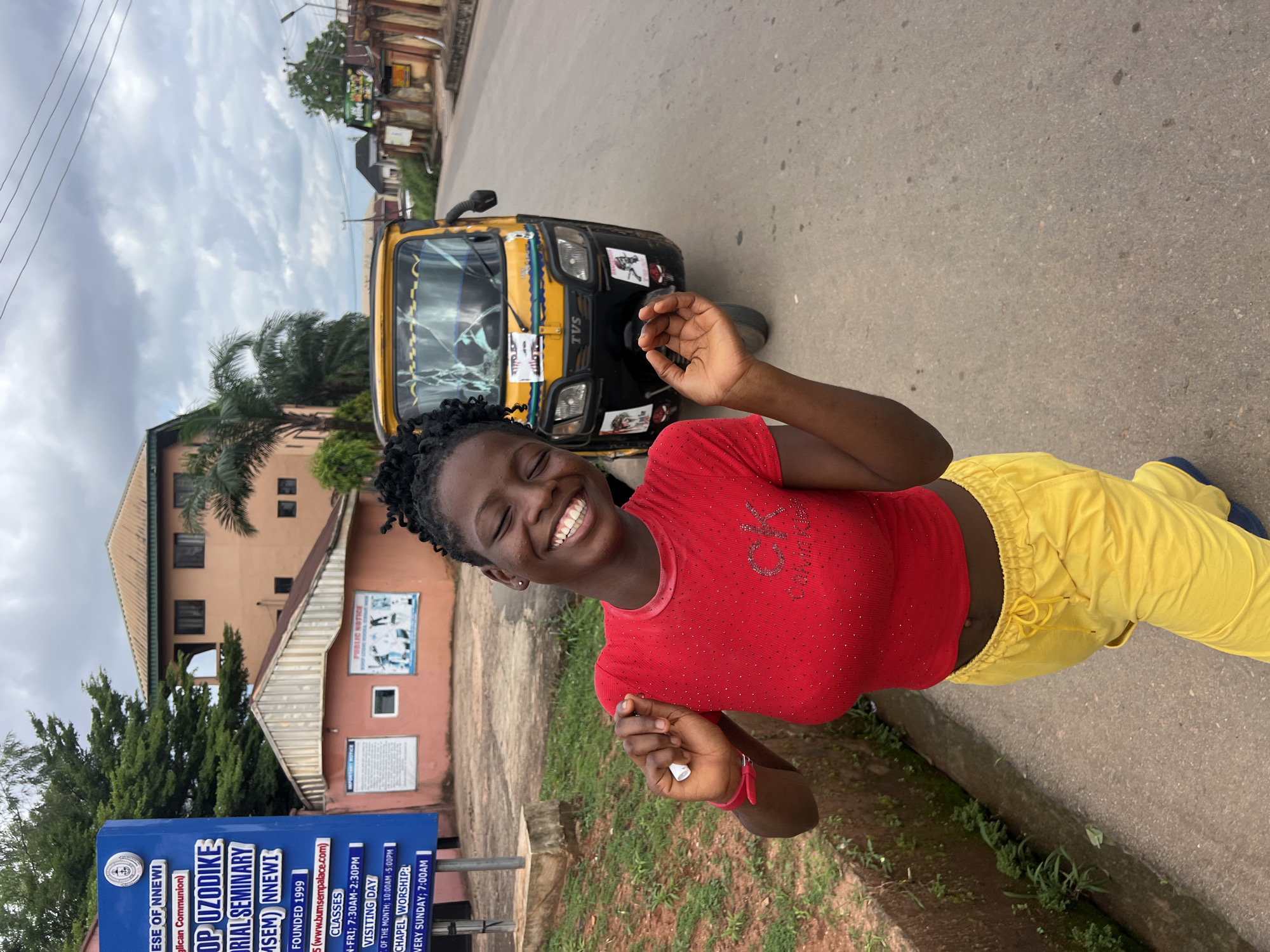 A Black female, appearing to be a teenager or young adult, smiles widely with her eyes closed as she poses for a photo in front of a yellow auto-rickshaw on a paved road. She is wearing a red, short-sleeved t-shirt adorned with rhinestones that form the letters "CK" and yellow drawstring pants. Her hair is styled in braids pulled back from her face.
To her left, a blue sign post is visible, with text that reads "DIOCESE OF NNEWI," "Bishop Uzodike Memorial Seminary (BUMSEM) NNEWI," "FOUNDED 1999," and information about classes and chapel worship. Behind the sign is a two-story building with a tiled roof and several windows, some of which are shaded. Adjacent to this building is a smaller, peach-colored structure with a peaked roof.
The yellow auto-rickshaw, a common mode of transport in Nigeria, has "TVS" written on its front and several stickers, including what appears to be a religious figure or image on the side. The road surface is asphalt, with a grassy and overgrown drainage ditch to the left of the woman. In the background, more buildings and vegetation are visible, suggesting an urban or semi-urban environment within the city of Nnewi, Nigeria. The sky is overcast with clouds.