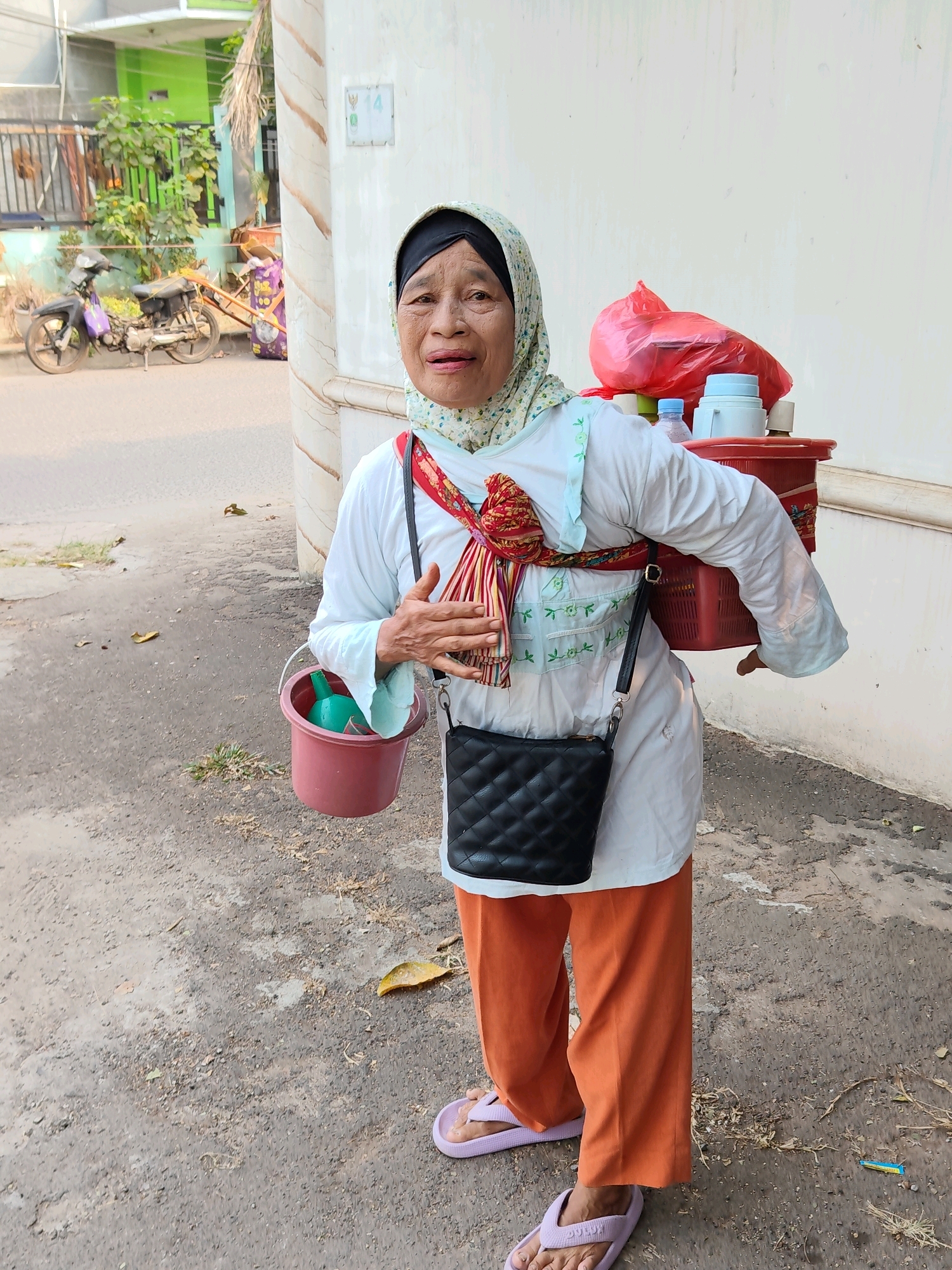 A street vendor, an elderly Indonesian woman wearing a headscarf, a white top, and orange pants, stands on a dirt and asphalt path in front of a cream-colored wall. She has a red basket filled with items and a red plastic bag on her back. A pink bucket with a green object inside hangs from her left side, and a black quilted purse is slung across her body. In the background, a motorcycle and a green building are visible, suggesting an urban environment in Bekasi, Indonesia. The lighting indicates it is daytime.