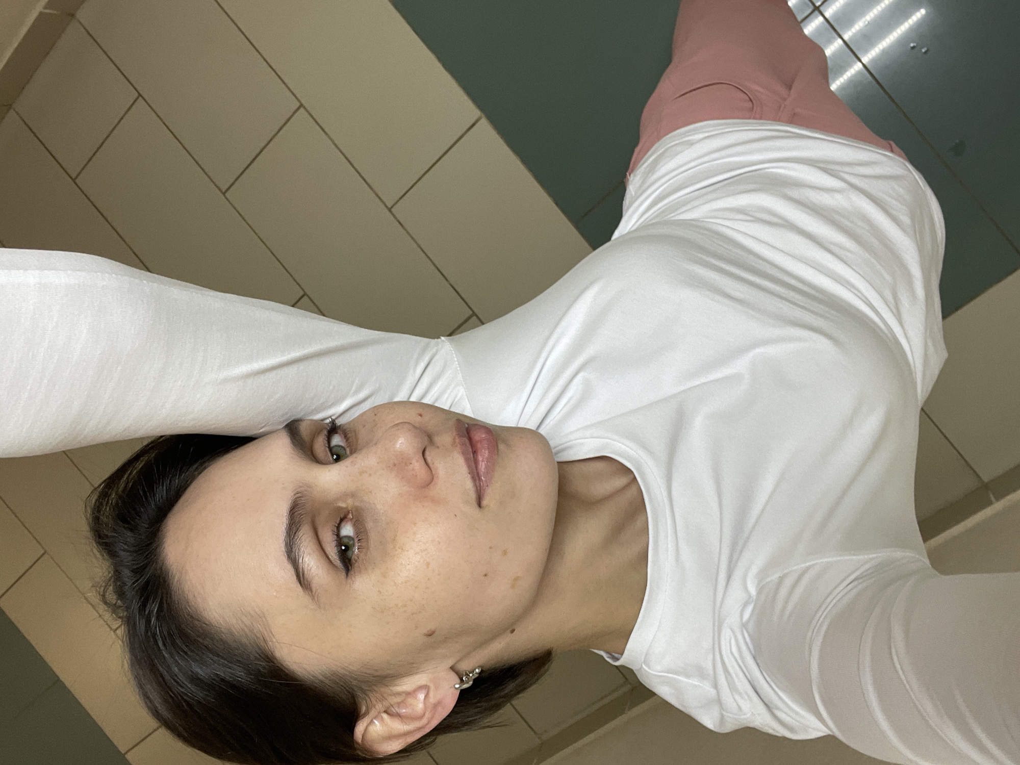 A young woman lies on a tiled floor, seemingly taking a selfie. She is wearing a white long-sleeved shirt and pink pants. The background includes light-colored and dark-colored tiles. Based on the tiles and lighting, the location may be a public space like a hallway, bathroom, or gym in Izhevsk, Russia. However, without additional context or specific landmarks, pinpointing the exact location in Izhevsk is not possible.