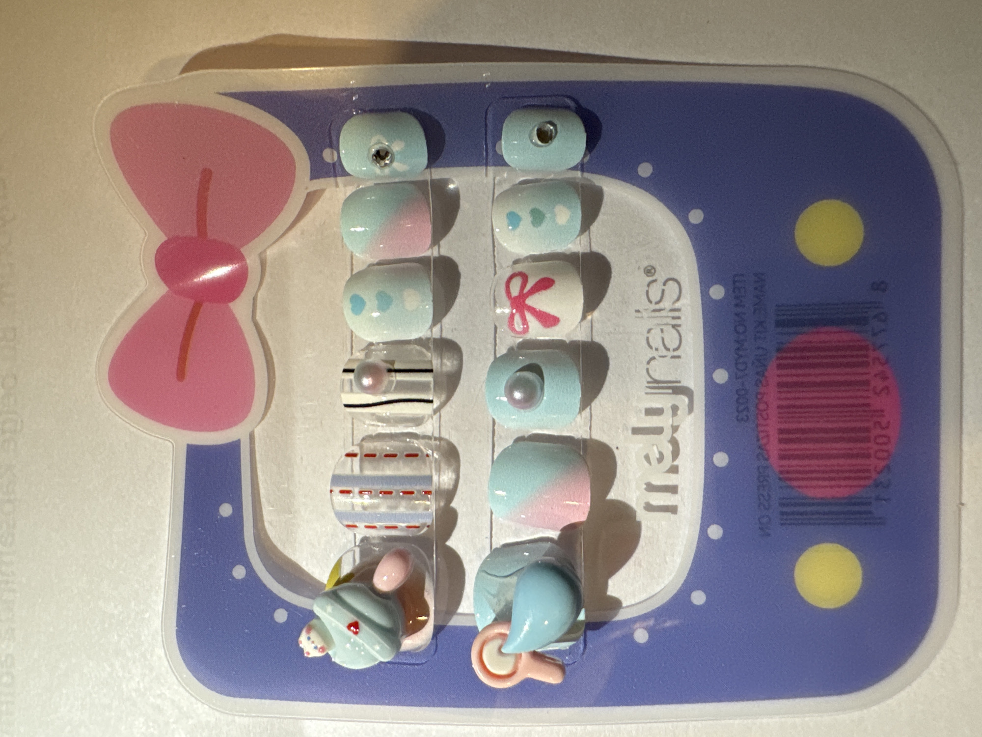 This image captures a package of colorful artificial press-on nails, likely intended for children or young adults, set against a light background. No people are visible.
The primary object is a blister pack containing a set of twelve unique false nails and two 3D decorative charms. The packaging features a large pink bow shape on the left, connected to a rectangular main display area colored in a light purple hue with white borders and scattered white dots. Two yellow circles are visible on the right side of the purple background.
The nails themselves showcase a variety of pastel designs, predominantly in light blue, pink, and white. Specific designs include:
* Solid light blue nails, some featuring a small rhinestone.
* Ombre nails, transitioning from light blue to pink.
* Light blue nails adorned with white and light blue heart patterns.
* White nails with a vibrant pink bow design.
* Transparent or white nails with thin black and red horizontal stripes, each featuring a small pearl.
* A light blue nail with a single pearl.
* Transparent or white nails with light blue and red horizontal stripes.
Additionally, two distinct 3D charms are included at the bottom: one appears to be a small light blue cupcake or hat with a pink heart detail, alongside a brown, possibly ice cream-like, element. The other charm is a light blue baby bottle or pacifier with a pink handle.
The scene is a static product shot, likely taken indoors on a flat surface, indicated by the shadow cast by the packaging. The lighting is artificial. There are no direct cues within the image to indicate the time of day, weather, or the provided location context of Buenos Aires, Argentina.
Visible text on the packaging, partially obscured and mirrored due to reflections, includes the brand name "melynails" (stylized), a registered trademark symbol "®", and reversed product information such as "ITEM NO.", "MADE IN", and what appears to be a barcode. Numbers like "8", "12005", and "SA2T1B" are also discernable from the reflected text.