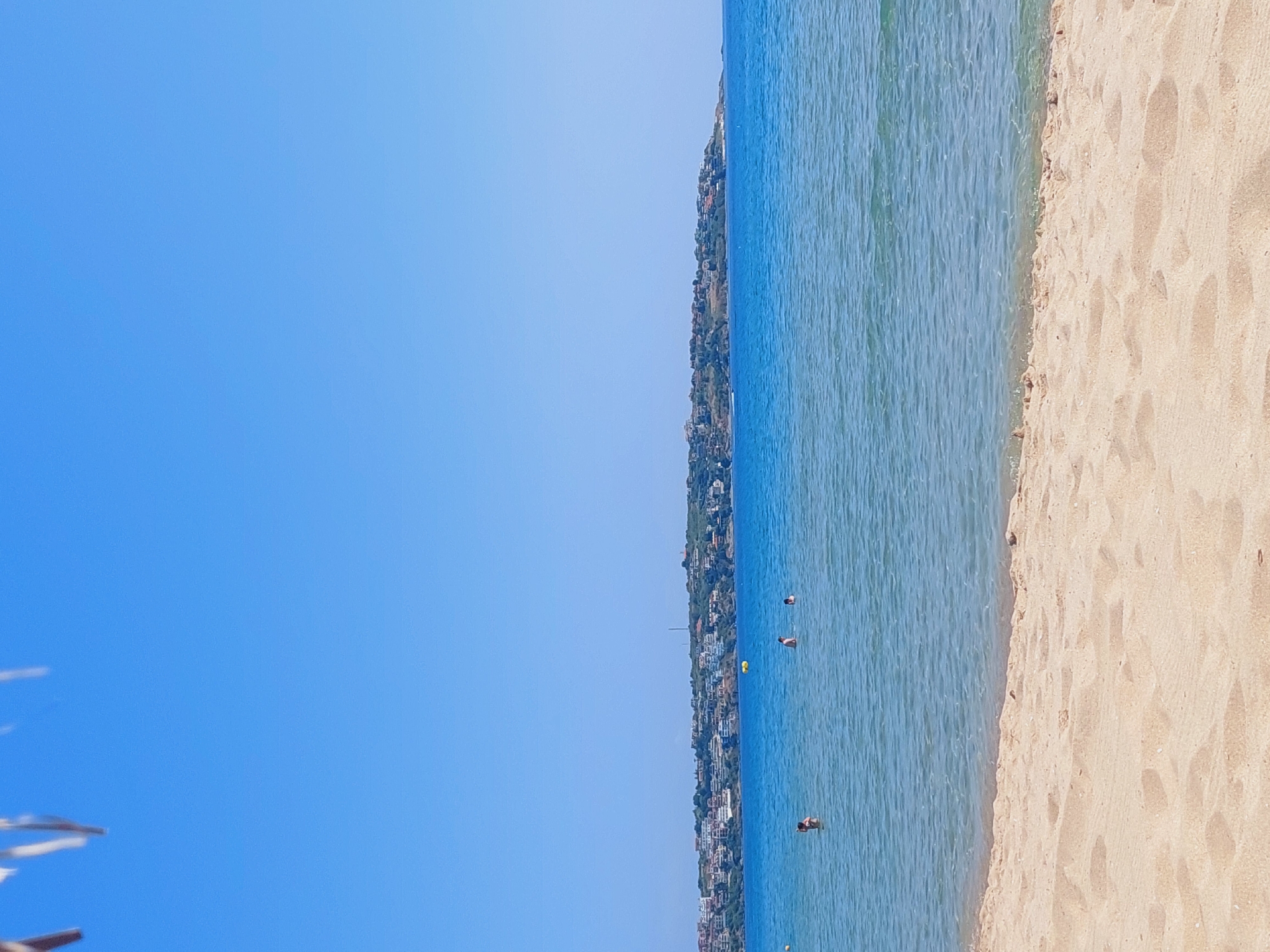 The image captures a tranquil coastal scene in Sozopol, Bulgaria, with a clear, azure sky dominating the upper portion. In the foreground, a sandy beach stretches out, its texture a tapestry of light and shadow. Gentle waves lap at the shore, creating a subtle froth where the water meets the sand.
Further out, the calm sea is populated by a few individuals swimming. One person is visible closer to the shore, appearing to be wading or swimming casually. Further out, two more individuals are present in the water, their bodies partially submerged. A yellow buoy floats near the center of the water, a solitary marker in the vast expanse.
In the distance, a picturesque coastline unfolds, characterized by a gentle slope dotted with numerous white and terracotta-roofed buildings. These structures cluster together, creating a charming hillside settlement. The greenery of trees and bushes is interspersed among the buildings, adding a natural contrast to the architecture. The overall impression is one of a serene summer day at a scenic seaside resort town.