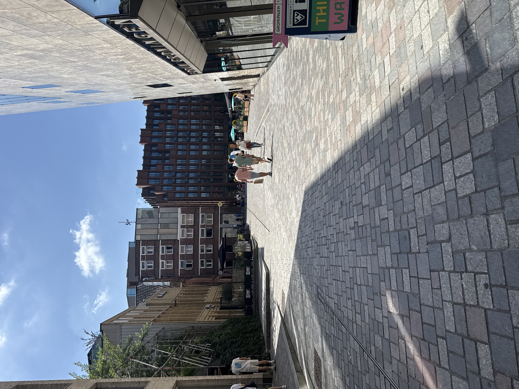 This is a street-level, vertical, full shot image taken in Groningen, Netherlands, on a sunny day. The scene depicts a cobblestone plaza or street lined with modern and older buildings.
On the left side of the image, a modern building with vertical wooden elements is visible, adjacent to a building with scaffolding and greenery. Further down the street, a multi-story brick building with numerous windows is present.
In the center of the image, several people are walking. Two men in the foreground are walking away from the camera, one in a pink t-shirt and the other in a light green t-shirt. A woman in a white top and jeans is walking in the same direction. Further back, a woman in a white top and a man in a light brown top are walking away from the camera.
On the right side of the image, a large, modern building with a textured concrete facade and a prominent sign that reads "FORUM GRONINGEN" is partially visible. A directional sign with the text "GRONINGEN" and "THIS WAY" in pink and green is positioned at the lower right corner.
The ground is paved with grey and pinkish cobblestones, with shadows cast by the buildings. The sky is blue with scattered white clouds. The overall impression is of a bustling urban environment on a pleasant day.