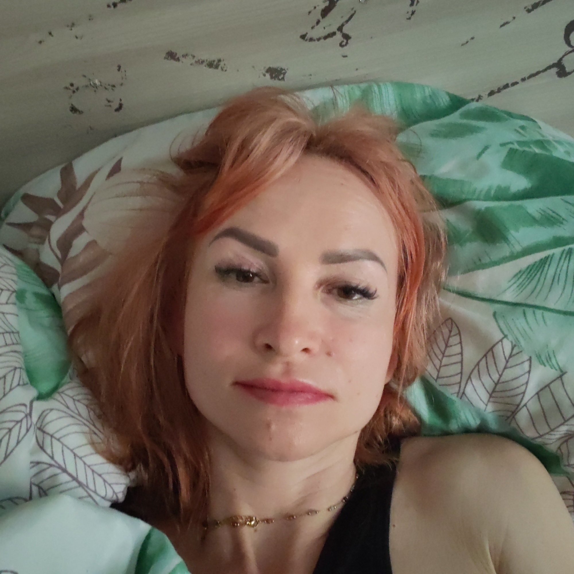 A woman with strawberry blonde hair is lying in bed. She is wearing a black top and a gold necklace. The bed has two pillows, one with a green and white leaf pattern and the other with a brown and white pattern. The wall behind the bed is light-colored with dark markings.
There is no indication that this image was taken in the city of Chaikovsky, Russia.