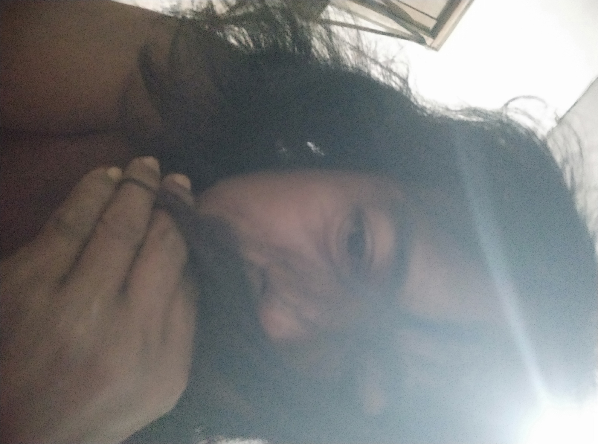 A close-up, slightly blurry, over-the-shoulder selfie shows a person with long, dark hair covering much of their face. The person's right hand is raised to their mouth, obscuring the lower half of their face. The lighting is bright and appears to be coming from the upper right, creating a halo effect around the head and washing out much of the background. The environment appears to be indoors, possibly a bedroom, with a window or door frame visible on the left side. The image's resolution and focus make it difficult to discern specific details of the person's features or the surroundings with certainty. There is no discernible information about the location in Sumurber, Indonesia.