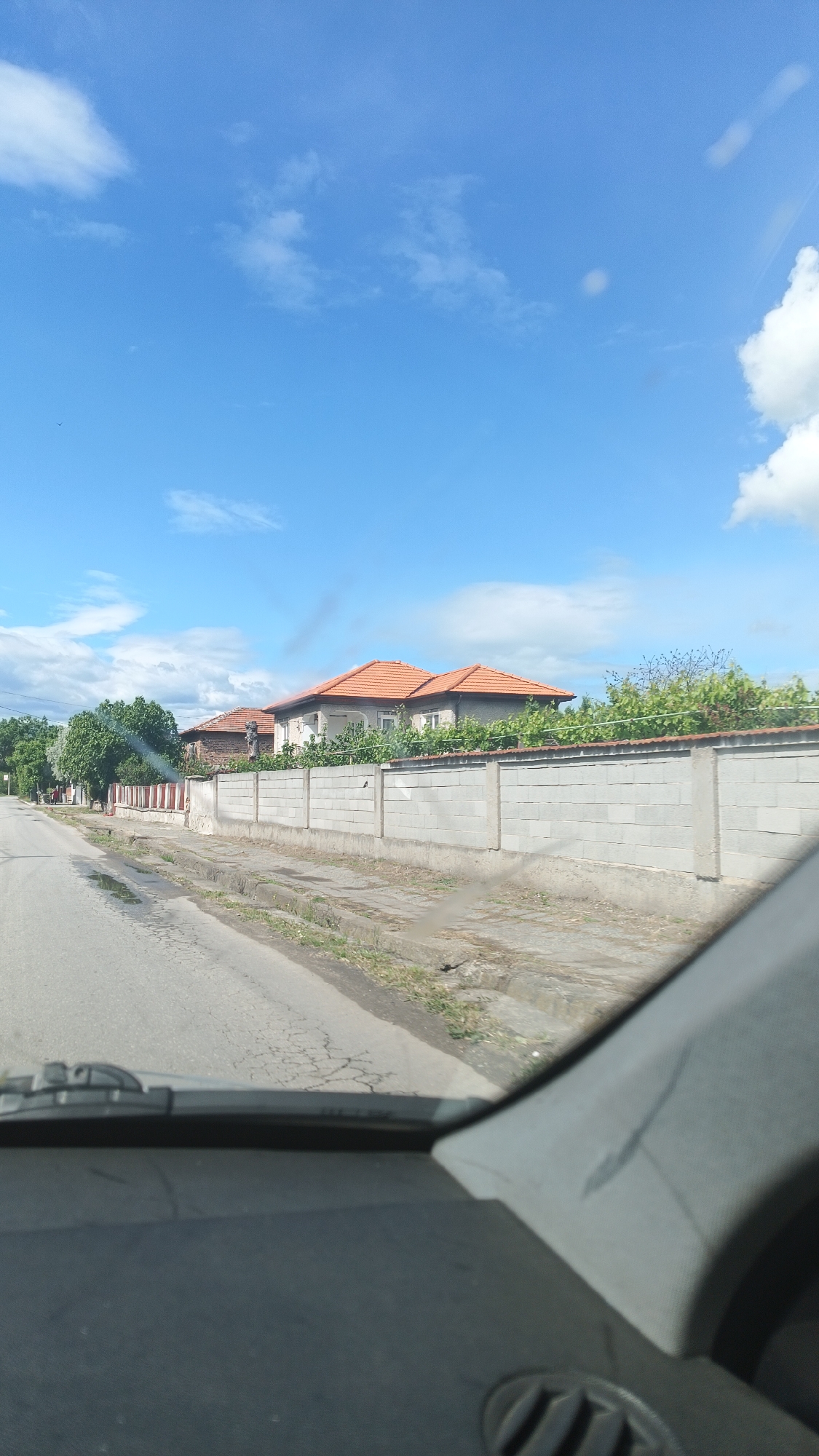 The image depicts a street in Boliartsi, Bulgaria, as seen from inside a vehicle. The road is paved with cracks and patches, and the view is obstructed by the car's dashboard and interior. On the right side of the road, a long, gray concrete block wall extends along the property line of a house with an orange tile roof. Vegetation is growing over the wall. In the distance, trees line the street. The sky above is partly cloudy, with blue visible.