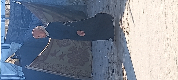 An individual in a black robe with a white collar and bald head stands near a structure covered with patterned fabric. The person appears to be in prayer or deep contemplation. The area around them seems to be a makeshift shelter or camp. The ground is sandy or dusty, and the lighting suggests an outdoor setting with strong sunlight casting shadows.