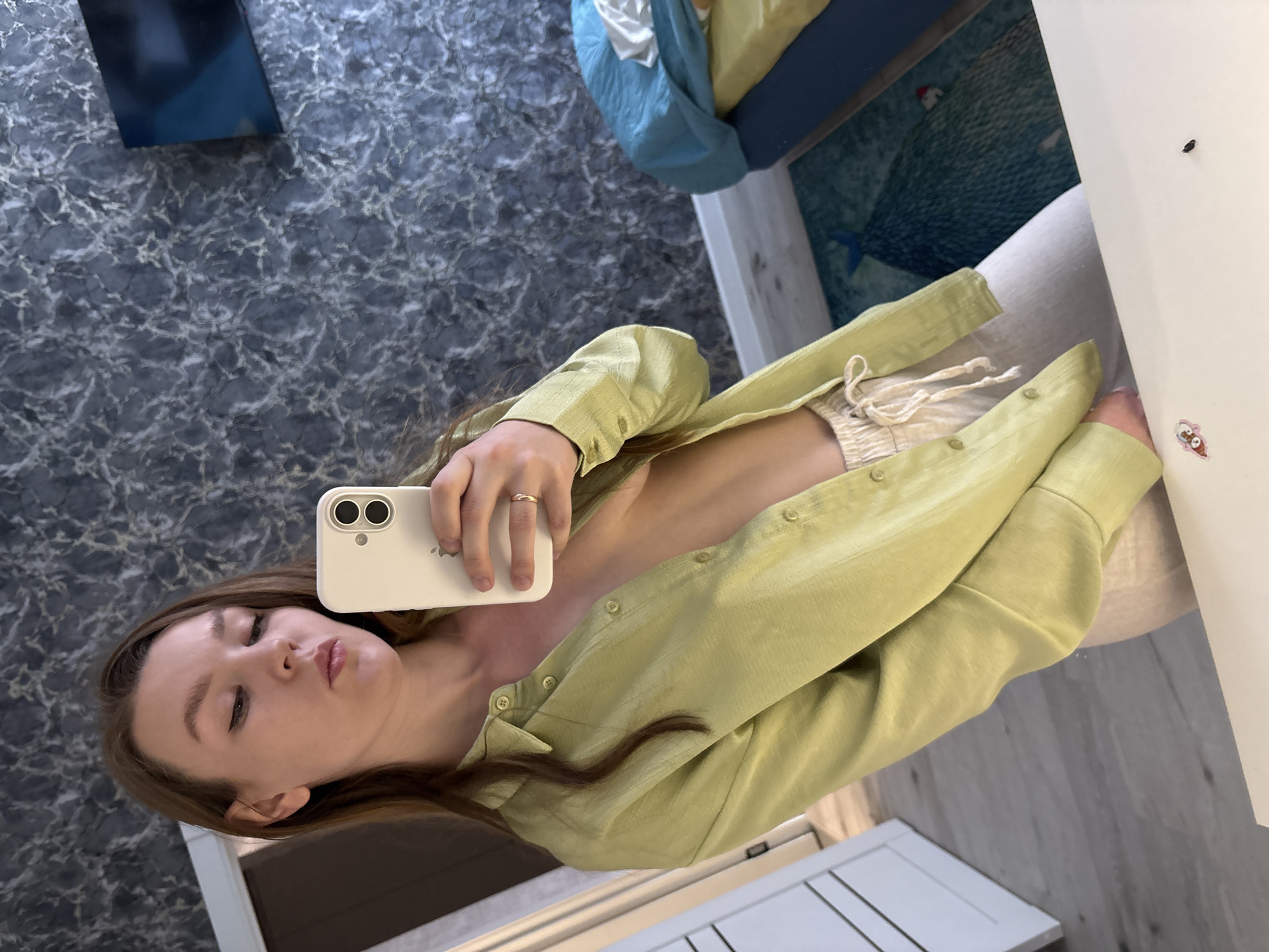 A woman takes a selfie indoors, wearing a light green button-down shirt and shorts. She is holding a white iPhone with a visible apple logo. The background includes a gray marble-patterned wall, a bed with blue and yellow bedding, a white desk with a cartoon sticker, and a white door. The specific location in Izhevsk, Russia cannot be determined from the image.