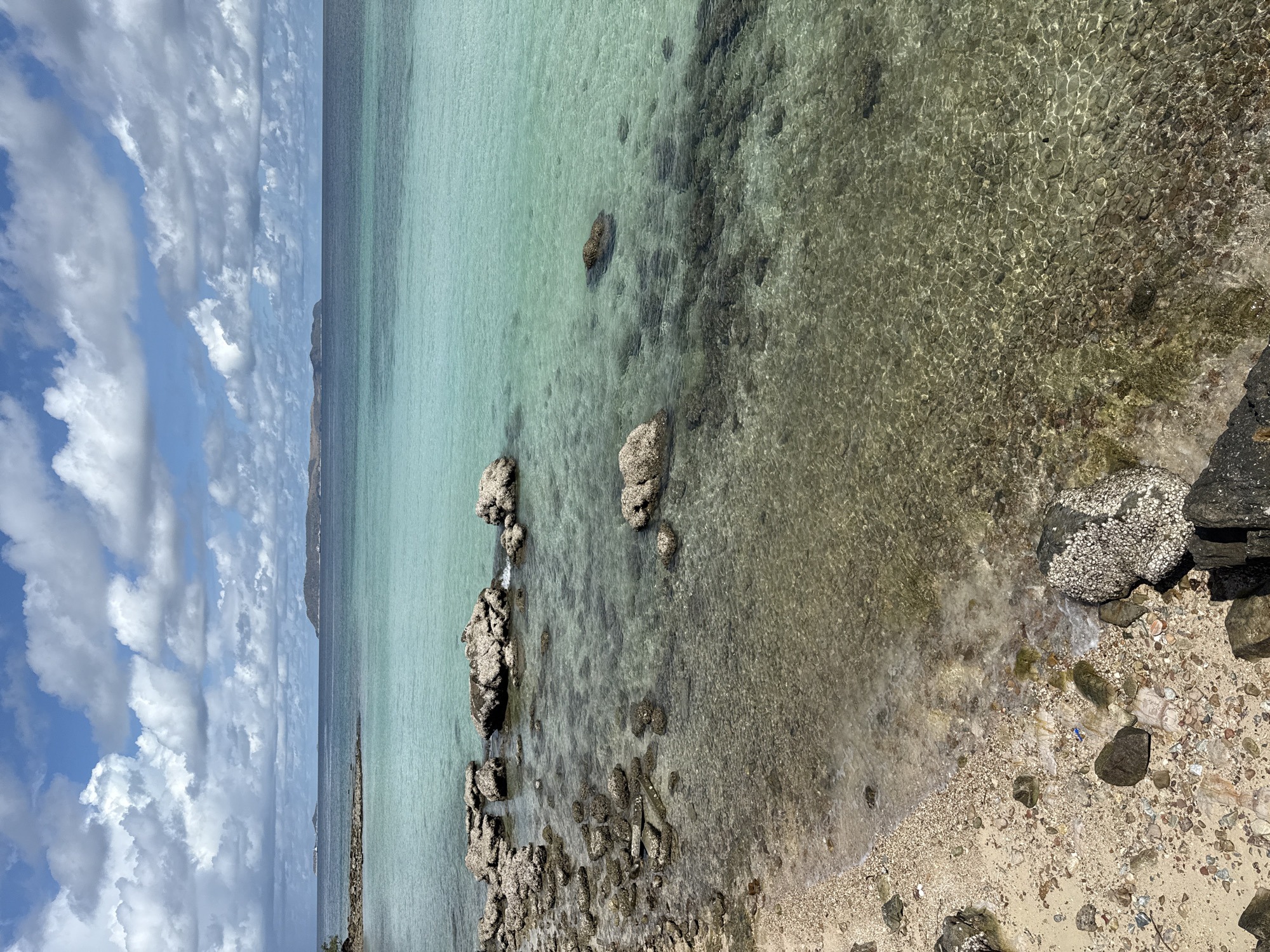 The image shows a coastal scene in Pattaya City, Thailand. The foreground features a sandy beach with scattered rocks and debris. Clear, shallow water extends from the beach, transitioning from a turquoise hue to a more transparent appearance, revealing the seabed. Rocks and coral formations are visible beneath the water's surface. In the distance, a coastline is discernible, and the sky is partially covered with white, puffy clouds.