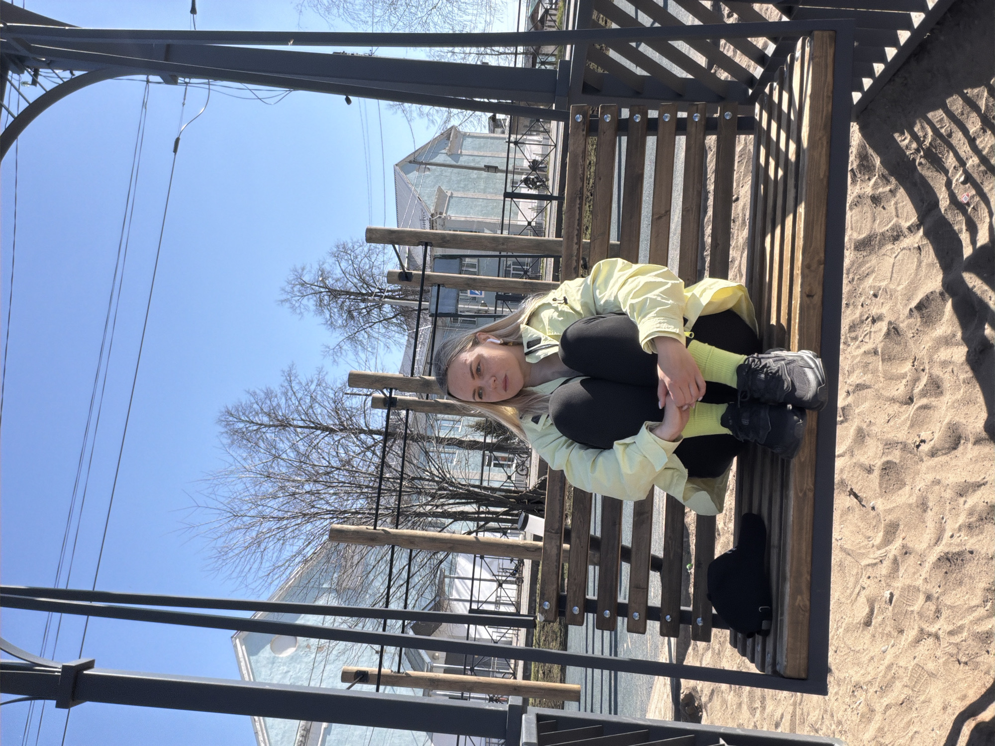 A young woman with blonde hair sits cross-legged on a wooden bench in what appears to be a public space. She wears black leggings, a light yellow jacket, and bright yellow socks with dark sneakers. Her face is visible, and she looks towards the camera. Behind her, a two-story building with light blue walls and a grey roof is visible through the bare trees. Above, a clear blue sky stretches overhead. The bench is part of a metal structure with wooden slats, casting shadows on the sandy ground beside the bench. A black bag rests on the bench next to her. The location is Votkinsk, Russia.