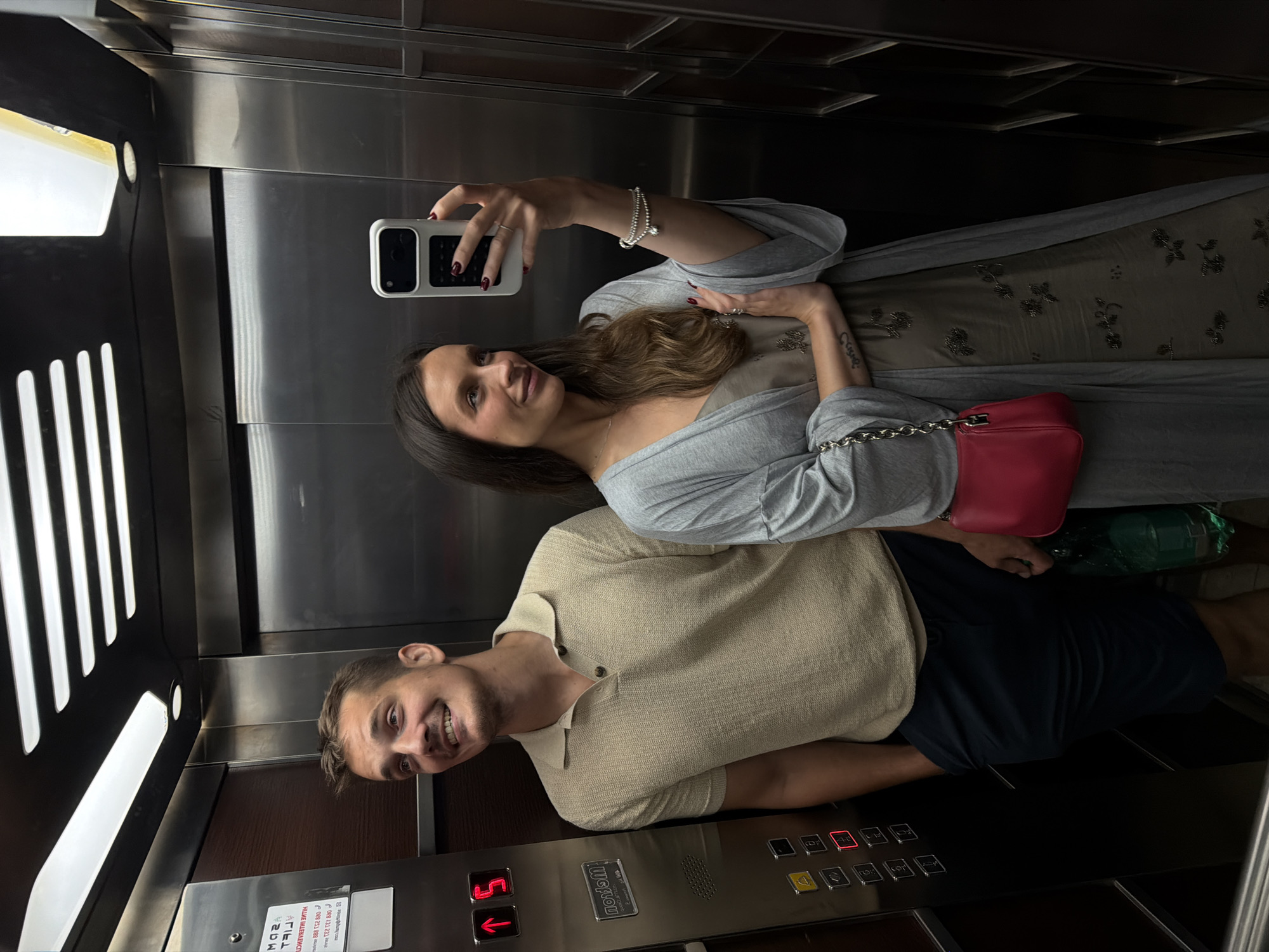 In an elevator, a woman holds a white phone and takes a selfie with a man. The woman has dark hair and wears a gray wrap. She also carries a red purse. The man has short brown hair and wears a tan shirt with dark blue shorts. They are both smiling. The elevator's control panel is visible with multiple buttons and indicators. The elevator's interior is metallic and wooden. The precise location within the city of Budva, Montenegro, cannot be determined from this image alone.