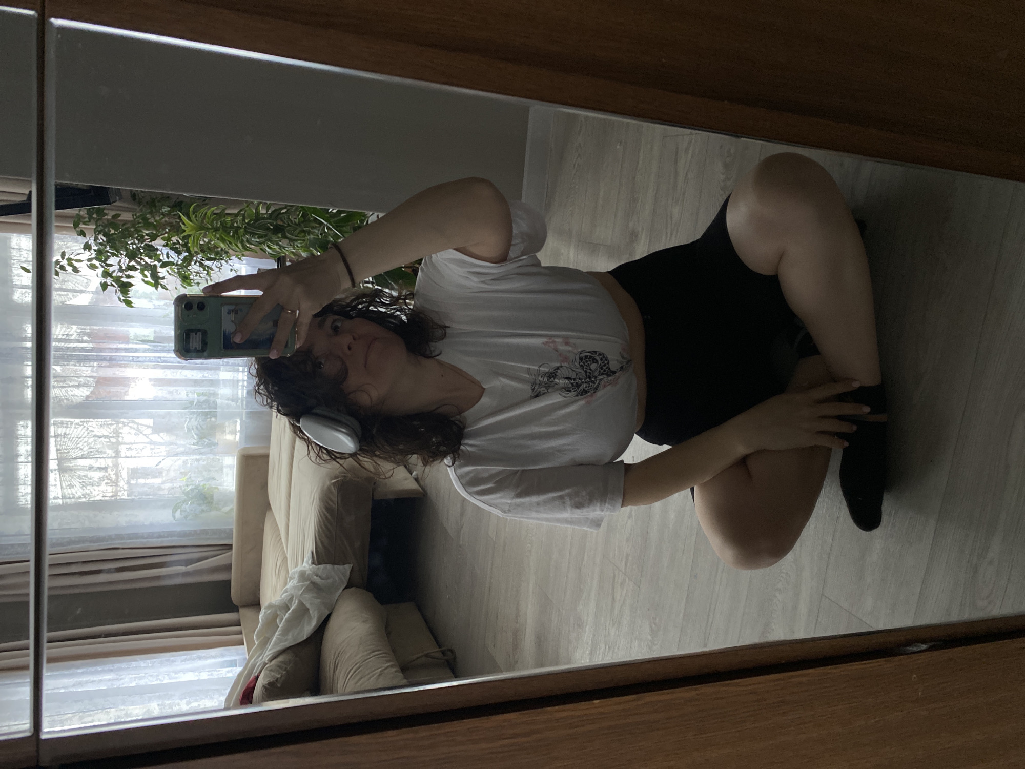 A reflection of a woman taking a selfie in front of a mirror. The woman is kneeling on a light-colored wooden floor, wearing a white t-shirt, black shorts, and a black sock on one foot. She holds a smartphone in her left hand, taking a photo of herself. The woman is wearing headphones. In the background, a beige couch and a window with sheer curtains can be seen. A plant is near the window. Location: Unknown, Unknown.