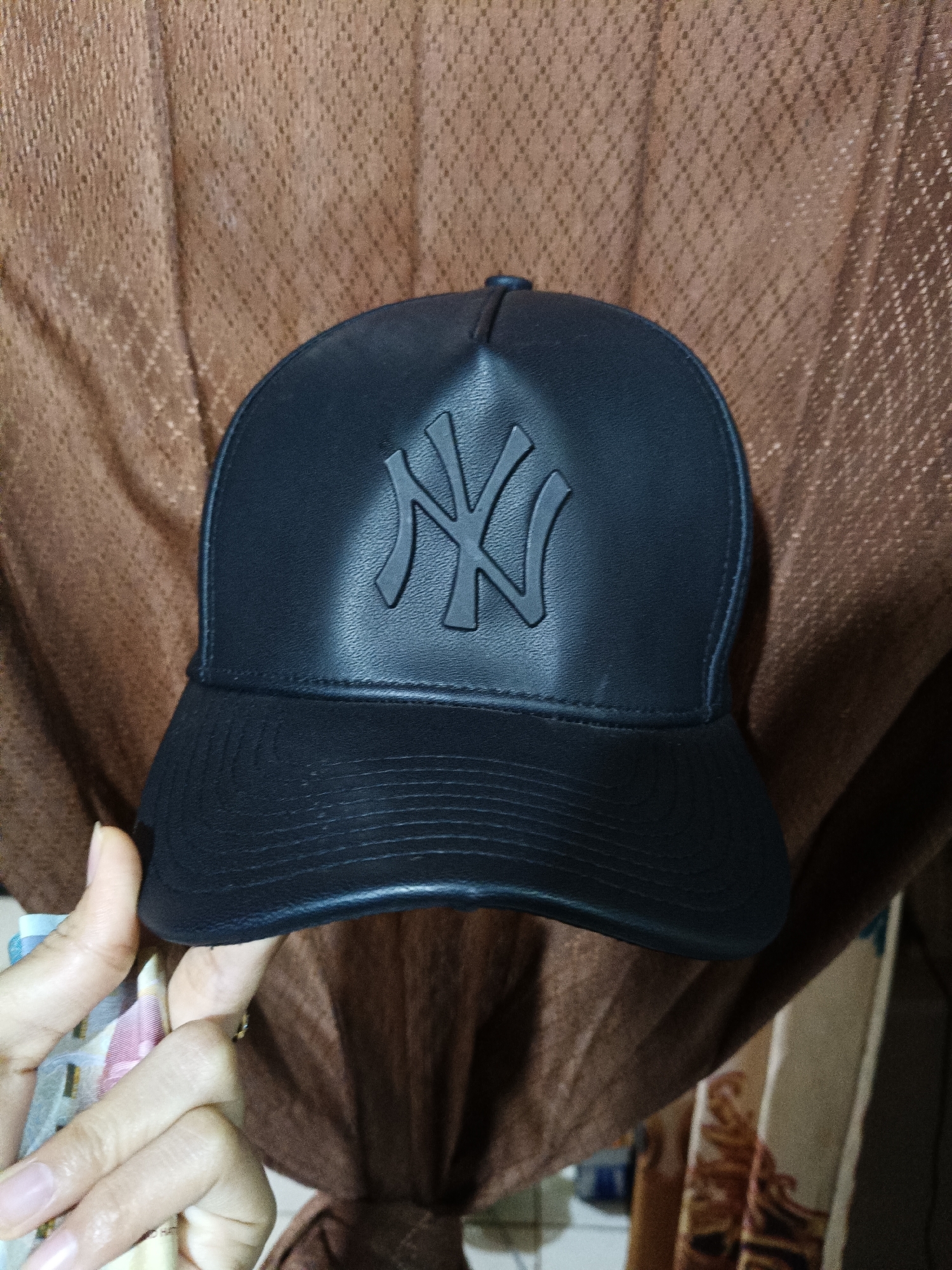 A black, faux leather baseball cap featuring a raised, monochromatic "NY" (New York Yankees) logo is centrally positioned in the frame, set against a draped, light brown fabric background. The cap has a structured crown and a flat brim with visible horizontal stitching lines. In the lower-left foreground, a hand, showing light to medium skin tone and natural nails, holds and partially fans out several banknotes, which display blue, pink, and cream colorations, consistent with Indonesian Rupiah. The background fabric exhibits a repeating diamond or lattice pattern. The scene is situated indoors, in Cileungsi, Indonesia.