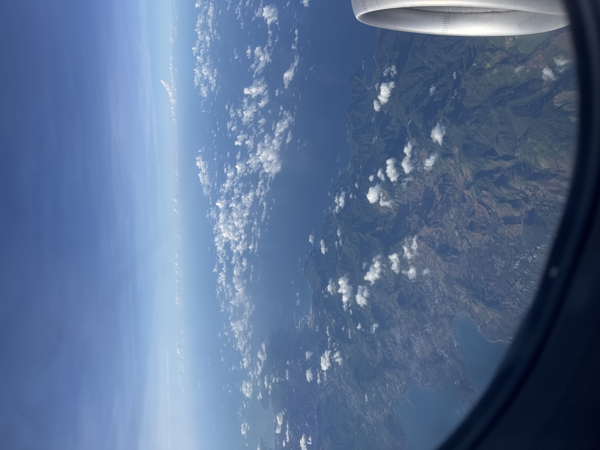 The image shows an aerial view from an airplane window. The sky is visible with scattered cumulus clouds. Below the clouds, the ocean or a large body of water meets a landmass, which appears to be mountainous and heavily forested with the sea near the coastline. Part of the airplane wing is visible at the top right of the image. The image does not contain enough information to determine the location.