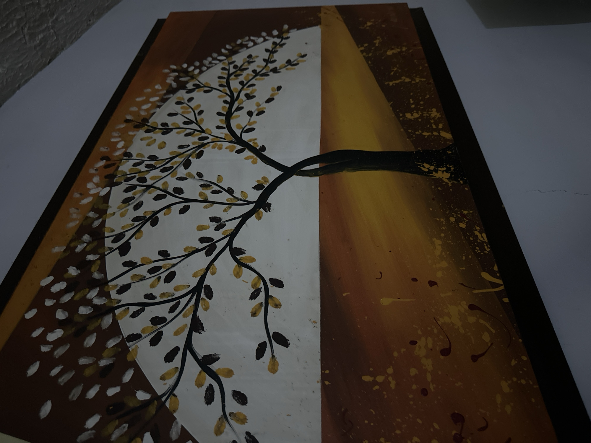 The image shows a rectangular painting with a dark brown frame, hanging on a light-colored wall. The painting is divided vertically into two distinct sections. The left section features a white background with a stylized tree. The tree has black branches adorned with leaves, rendered in a mix of gold and black. The leaves appear to be partially transparent. The right section has a brown background with yellow and darker brown splatters. A gradient of yellow is also visible. The tree trunk, depicted in dark brown and black, extends from the right section into the left. The photograph appears to be taken indoors under dim lighting conditions. There is no evidence to suggest the location is in Parroquia Ocumare, Venezuela.