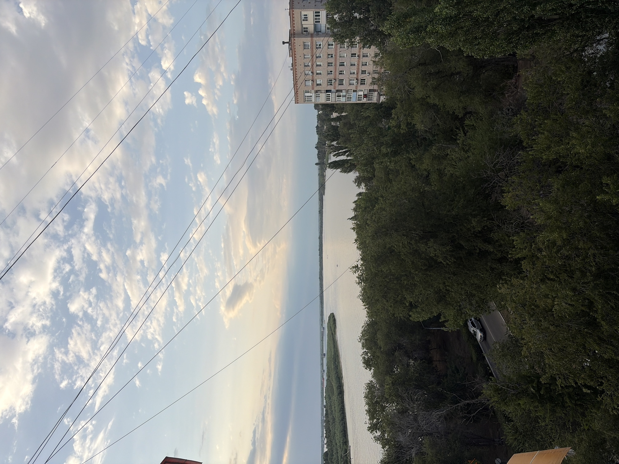 This is an exterior, eye-level, medium shot of a river and its surroundings in Astrakhan, Russia. The image is captured from a high vantage point, looking down and across a wide, calm river. In the foreground, a dense canopy of green trees dominates the bottom half of the frame, with a road running through them and a silver SUV driving on it. To the right, a multi-story apartment building, likely constructed from brick, rises above the trees. In the distance, the river stretches towards the horizon, with landmasses dotted with trees on its far bank. The sky above is a mix of blue with scattered white clouds, suggesting either early morning or late afternoon. Power lines are visible diagonally across the top of the image. The overall scene depicts a semi-urban environment bordering a natural waterway.