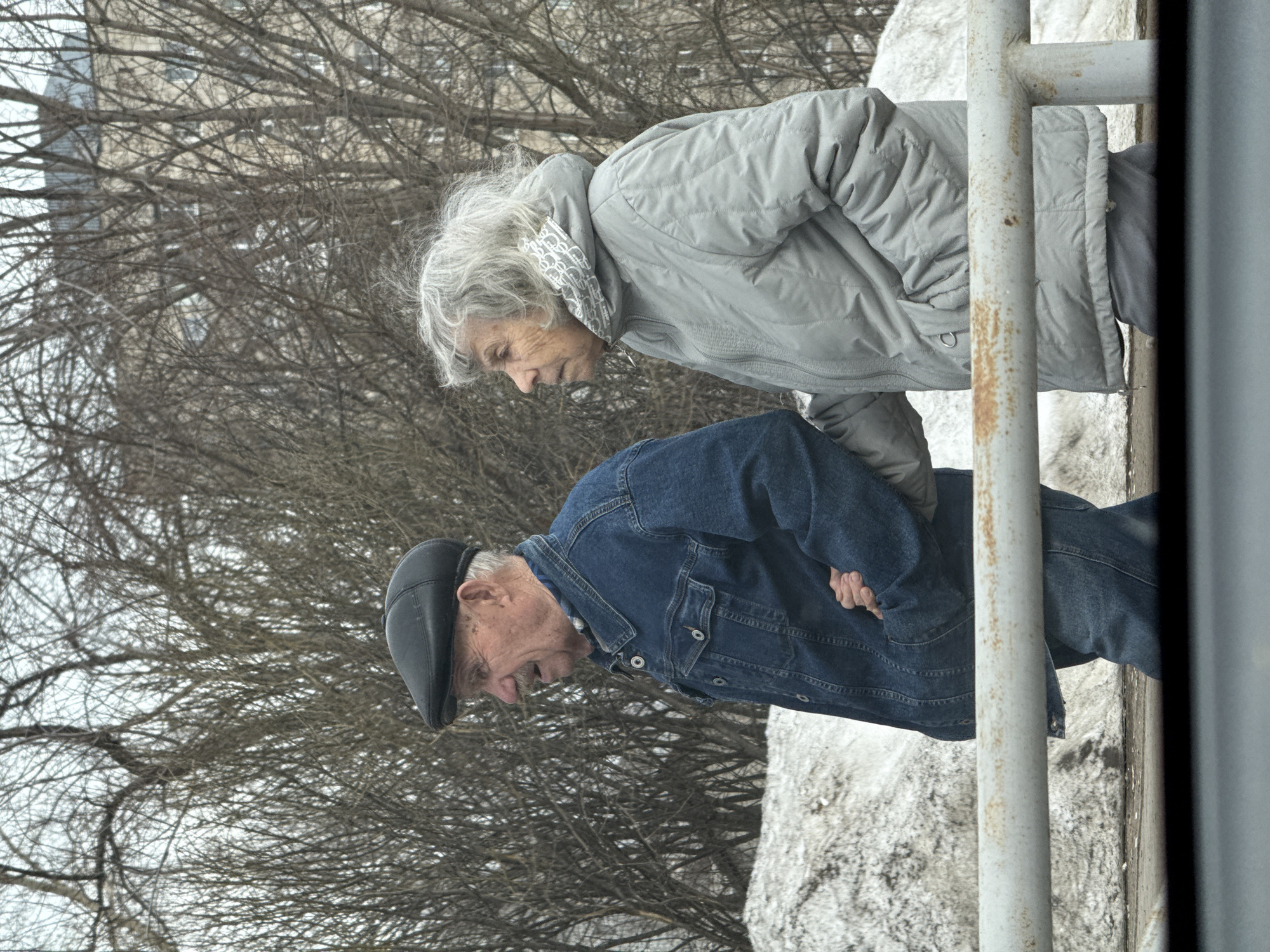 Two elderly individuals stand near a snow-covered area, with bare trees in the background. The woman, with gray hair, wears a light gray quilted jacket. The man, wearing a black cap and denim jacket, stands to the left of the woman. A gray metal railing is visible on the right side of the frame. This scene appears to be taken in Chaikovsky, Russia.