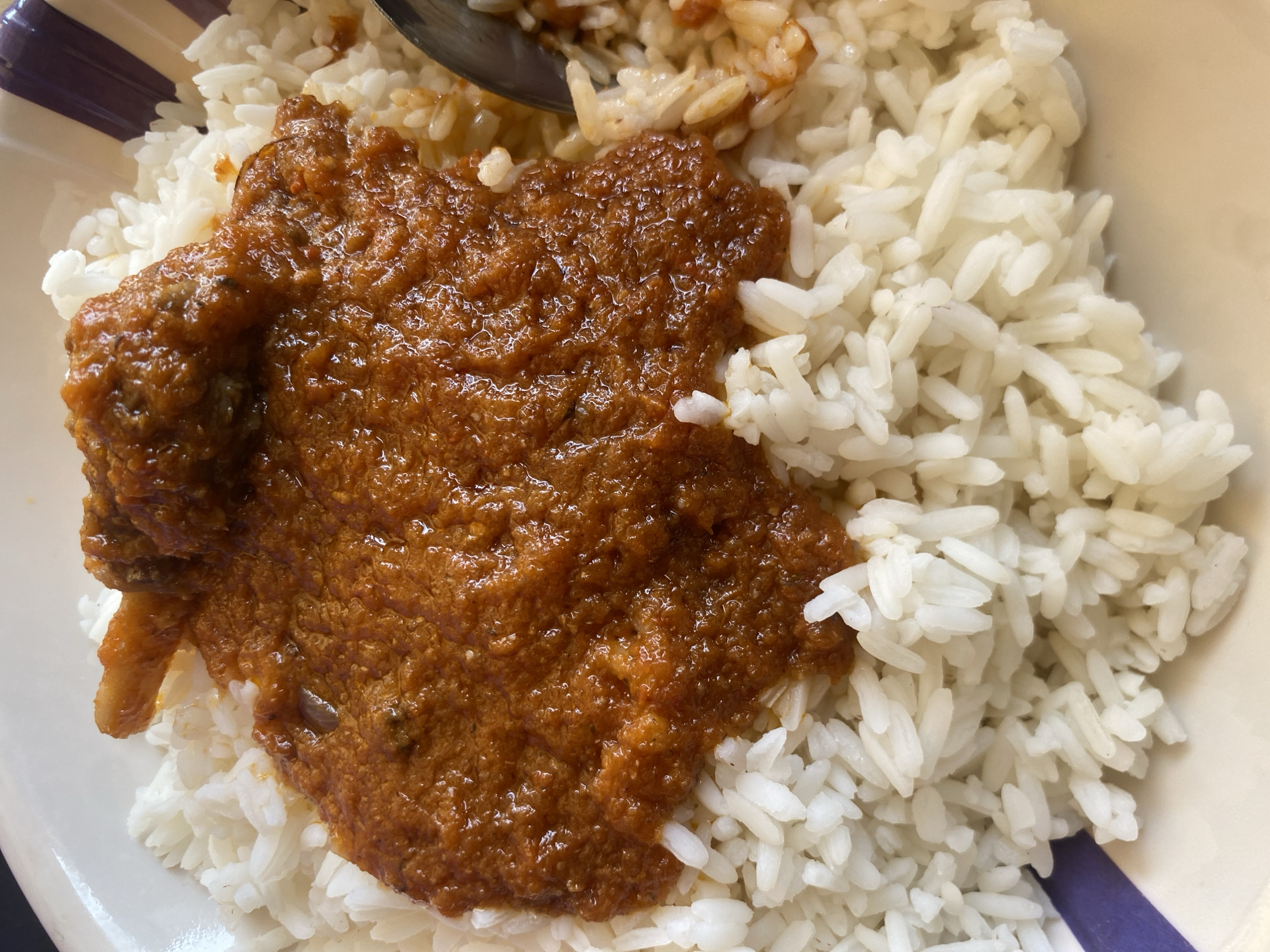 A top-down close-up shot shows a serving of white rice in a bowl, topped with a reddish-brown stew or sauce. A spoon is visible at the top of the frame, partially obscured by the rice. The bowl has white and purple stripes along its rim. The location is presumed to be Barden Yana, Nigeria, though this cannot be definitively determined from the image alone. No people are visible.