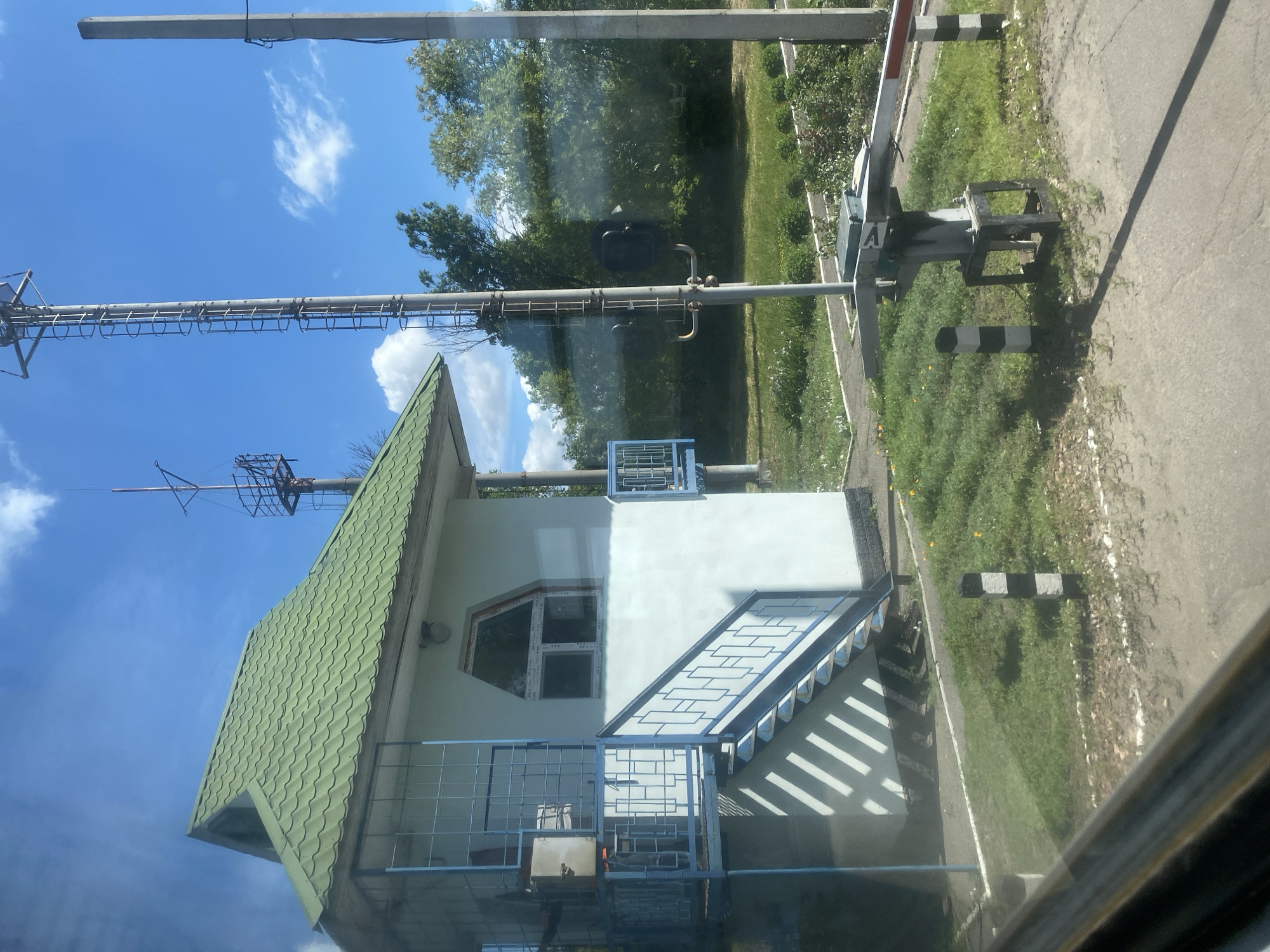 The image shows a two-story building with a green tiled roof, a light-colored facade, and blue metal railings. A tall metal tower with antenna-like structures rises from behind the building. A utility pole with horizontal supports and wires is visible in the upper left of the frame. A railroad crossing gate is partially visible in the upper right of the image, with black and white striped posts alongside a road. The sky is blue with some clouds. The location is Pidluky, Ukraine. The view is through a window, as evidenced by reflections in the foreground.