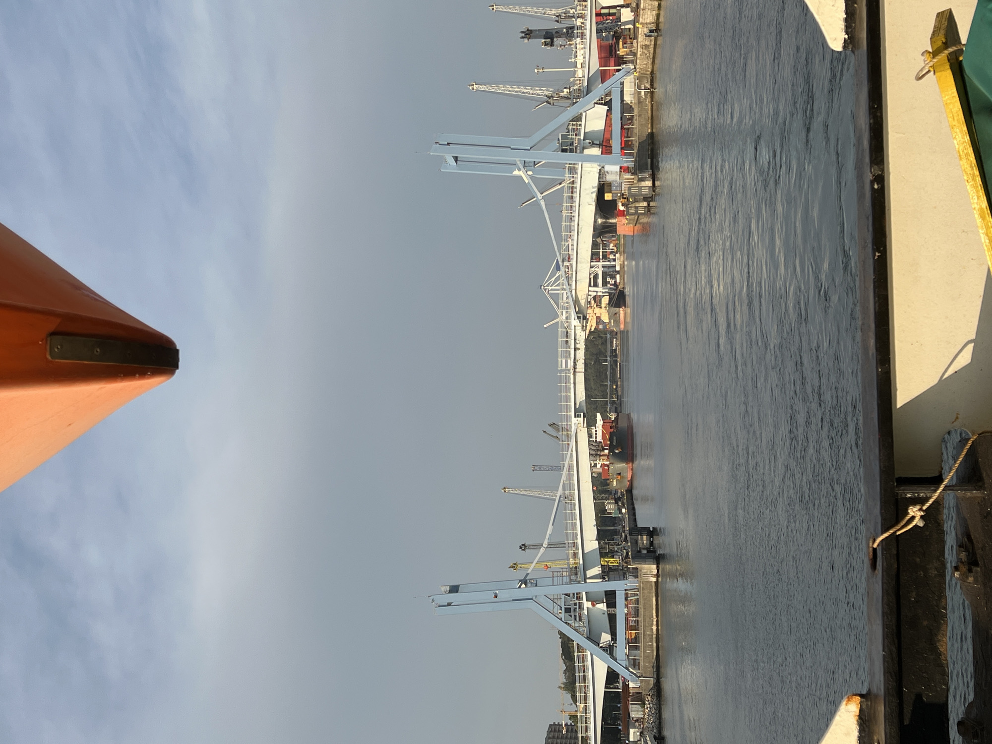 The image shows a view from a boat looking towards a drawbridge over a waterway. The drawbridge has two large blue towers and a white deck. There are several large ships and cranes visible in the background, suggesting a port or industrial area. The sky is overcast. The foreground of the image shows the edge of the boat the picture is taken from, with a rope tied to a cleat. The location is identified as Matosinhos, Portugal.