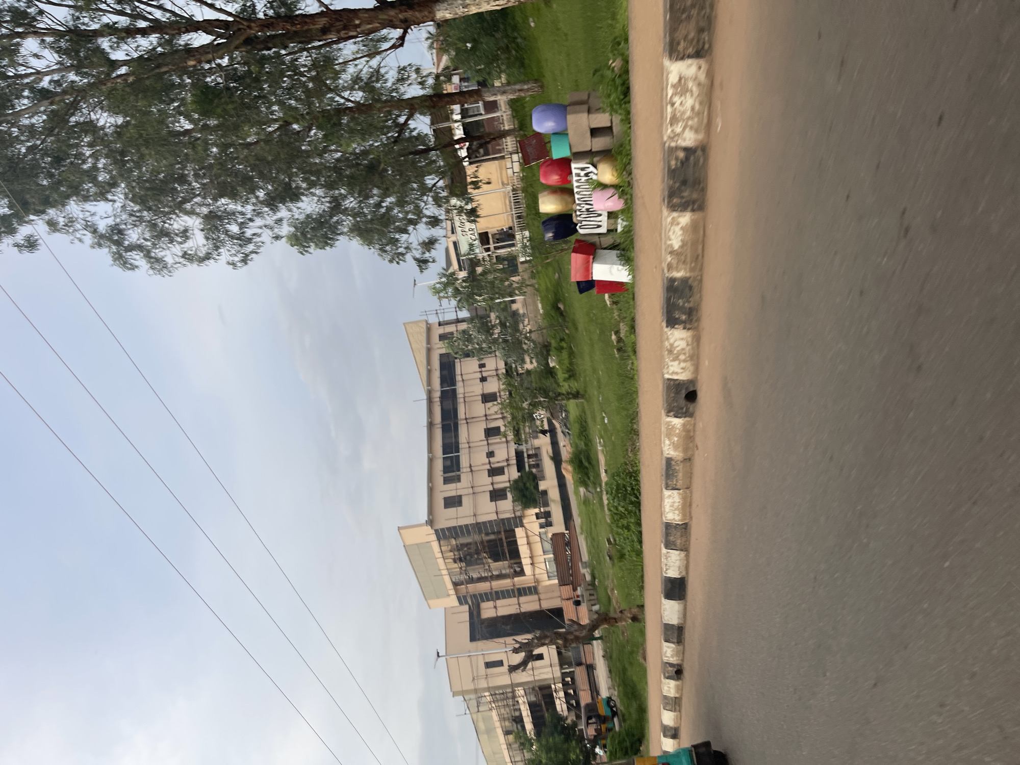 The image captures a street scene in Bar Jirgi Summa, Nigeria, with a multi-story building under construction in the background. The building is largely light brown and beige, with scaffolding covering parts of its exterior, suggesting ongoing renovation or development. In the middle ground, a grassy area is visible, dotted with trees. A collection of colorful, round pots and some white rectangular objects are arranged on a low wall in front of the trees, with a sign displaying the number "07062000654" among them. To the left of the building, a three-wheeled vehicle, possibly a rickshaw or auto-rickshaw, is partially visible. The foreground shows a paved road with a black and white striped curb. The sky is overcast with a light blue hue. Power lines are visible in the upper portion of the image.
