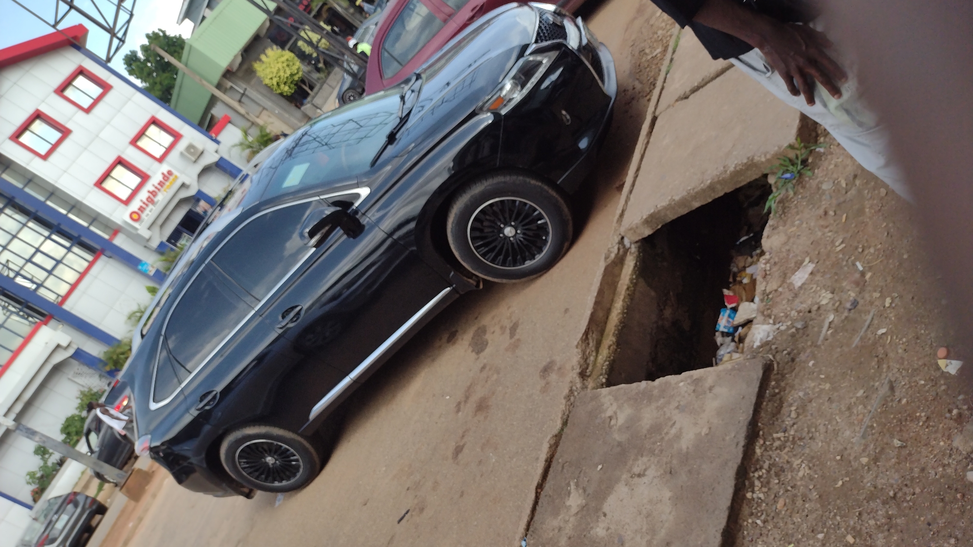 In Jos, Nigeria, a black Lexus SUV is parked on a dirt road next to a drainage ditch filled with debris. The car's windows are tinted, and it has black rims. To the left of the car is a modern, multi-story building with white walls, red accents around the windows, and blue trim. The building has the text "Onigbinde" in yellow lettering above a smaller sign. The scene appears to be set during the daytime, with the sun casting shadows. The overall impression is one of urban, possibly commercial, activity in Nigeria, with a juxtaposition of modern architecture and a less-than-pristine street environment. The image is taken at an angle, suggesting the photographer is standing to the side of the car.