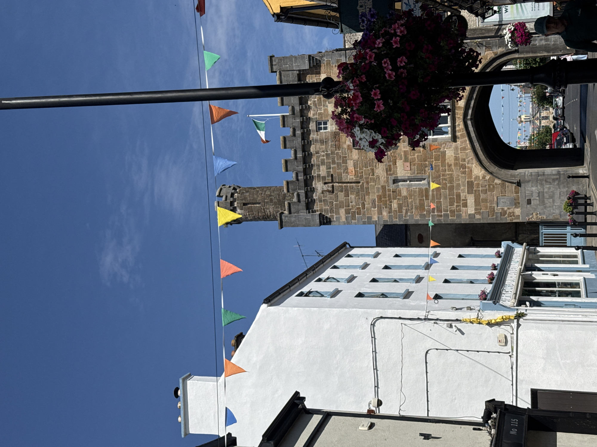A street view in Clonmel, Ireland, shows a historic stone archway with a tower on the right side of the image. An Irish flag flies from the top of the tower. Bunting in various colors is strung across the street, attached to a lamp post. A white building with blue-trimmed windows is on the left, with a black-trimmed awning over a shop entrance that reads "No 115". A hanging basket overflowing with pink and white flowers is suspended from the lamp post in front of the archway. The archway leads to a street with cars parked and driving, and more colorful bunting visible in the distance. A man wearing a green cap is partially visible on the right side of the image, facing away from the camera. The sky is clear and blue with a few scattered clouds.