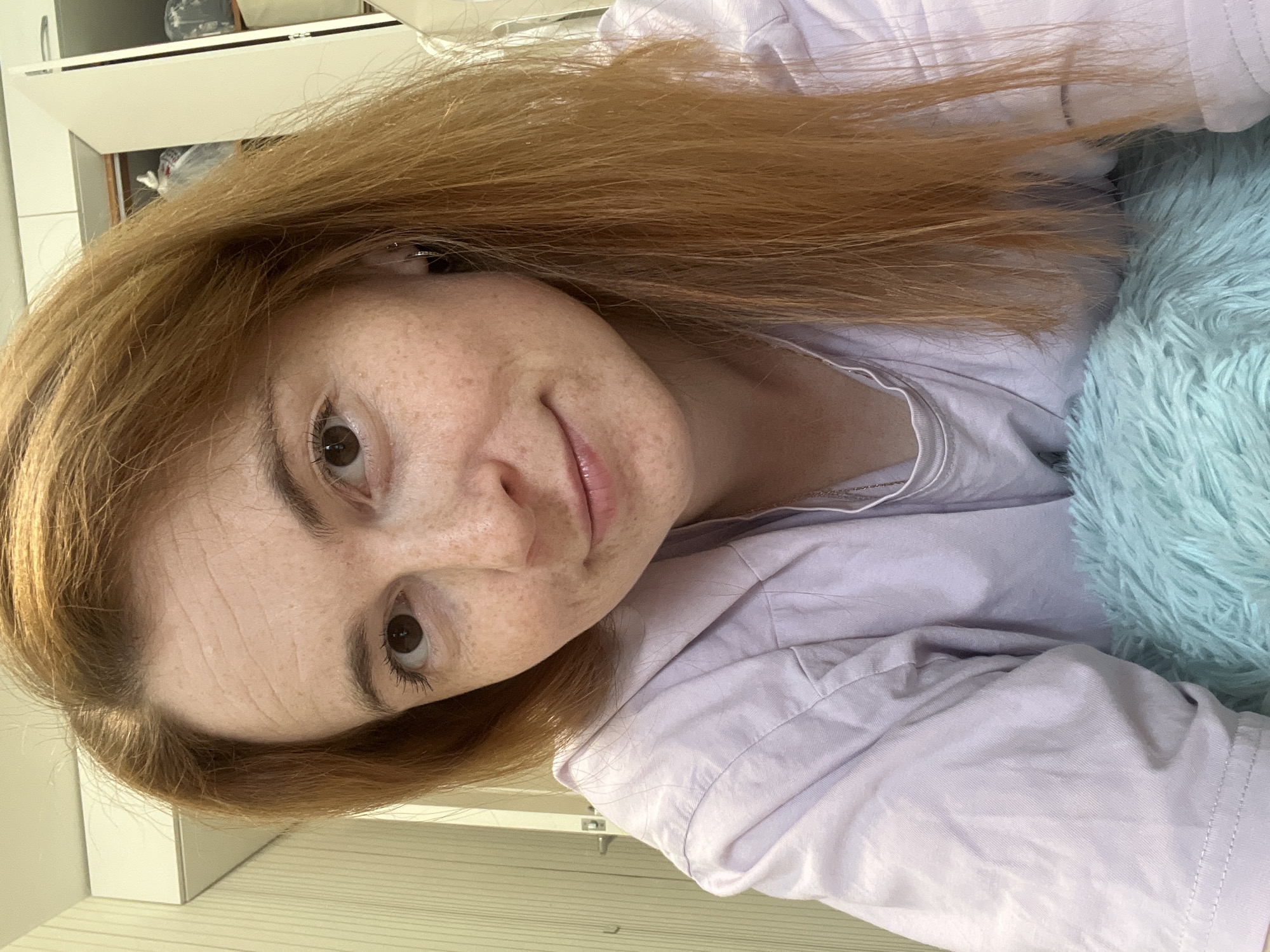 A fair-skinned woman with long, reddish-brown hair and numerous freckles on her face is captured in a close-up, eye-level portrait. She has brown eyes, minimal eye makeup, and a subtle smile. She wears a light lavender, collared shirt, possibly over a lighter undergarment, and a thin silver chain is visible at her neckline. A small silver hoop earring adorns her right ear. In the lower portion of the frame, a fuzzy, teal-colored textile, likely a blanket or pillow, is partially visible. The immediate background on the left consists of a light-colored wall with vertical striped paneling and a door handle. On the right, white overhead cabinets are open, revealing assorted items stored within. The setting appears to be an indoor, domestic environment.