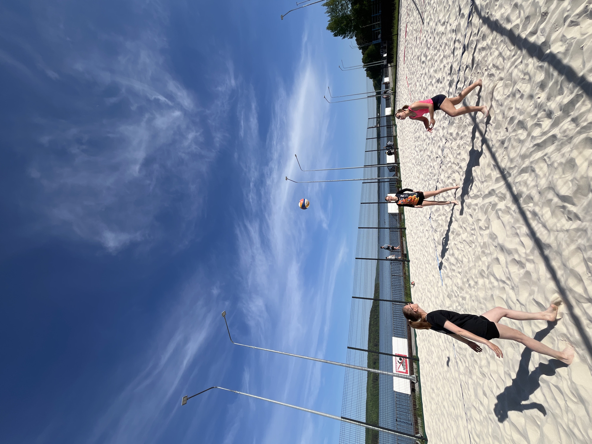 The image depicts a beach volleyball scene. Three young women are positioned on a sand court. One is wearing a black shirt and shorts, another a pink top with dark shorts, and the third a black and orange top with black shorts. A volleyball is suspended in the air between them. A chain-link fence runs along the edge of the court, with a body of water and trees visible beyond. Tall light poles are spaced along the side of the court. The sky is bright blue with streaky white clouds. The image was likely taken in the city of Chaikovsky, Russia, as indicated by the geographical details implied in the prompt.
