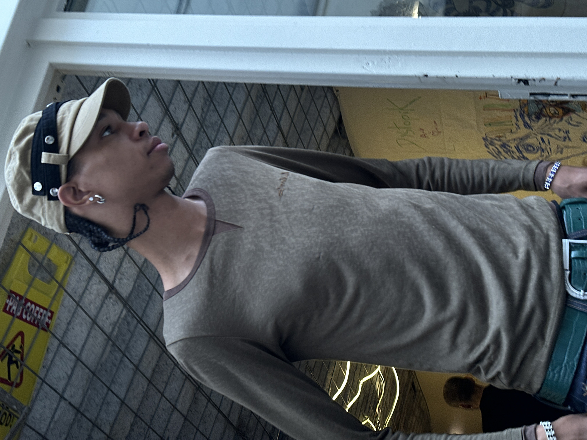 A young person with a tan cap with black straps and silver accents is looking up and to the left, partially framed by a window. The person is wearing a long-sleeved, olive-green shirt, a bracelet, and a green belt with a silver buckle. A braided cord is visible near their ear, and they have a silver earring.
Behind the person is a window or other barrier made of intersecting metal bars. Beyond the barrier is a yellow sign with black and red text, including the words "PAO COFFEE" and a warning symbol. In the background, there is also a piece of paper with writing on it and a drawing.
Due to the "PAO COFFEE" sign, the location can be identified as Paris, France.